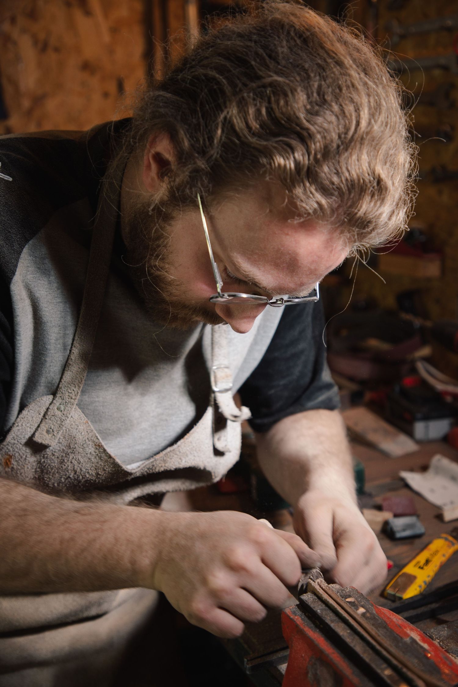 Bearded craftsman wearing safety glasses carefully working on knife blade at rustic wooden workbench.