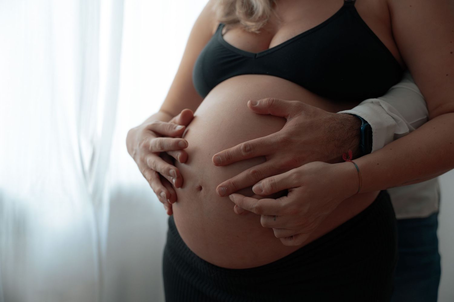 Close-up of pregnant belly with couple gently cradling bump with both hands indoors.