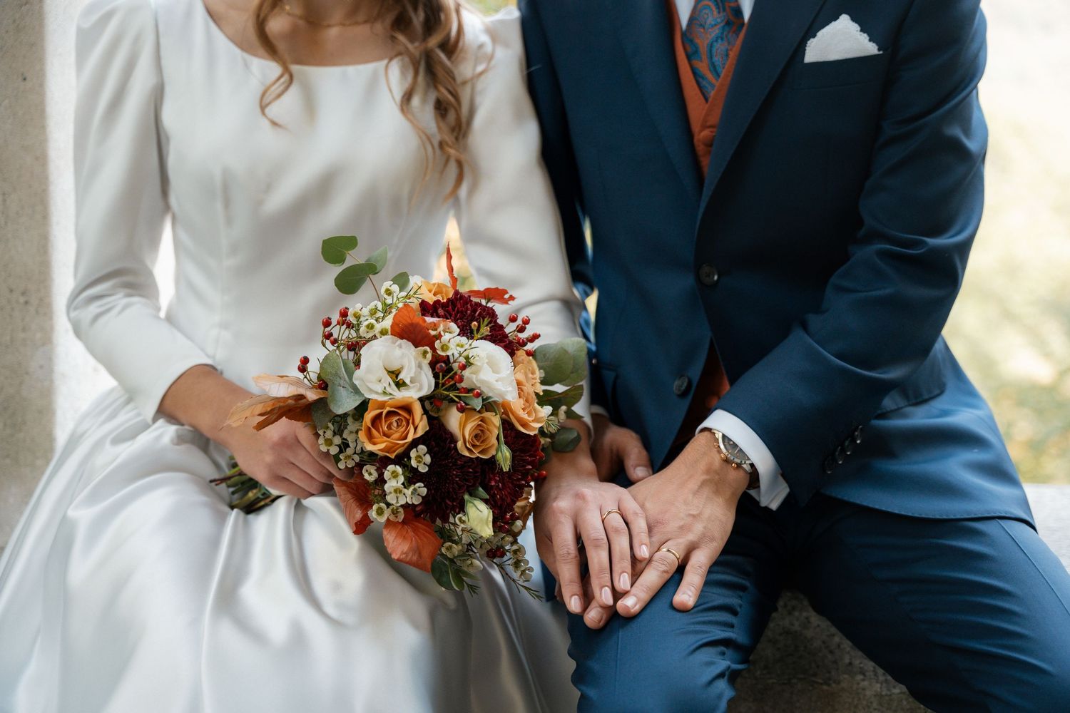 Bride and groom sitting together, showcasing wedding rings, bridal bouquet with autumn flowers in orange, red, and white.