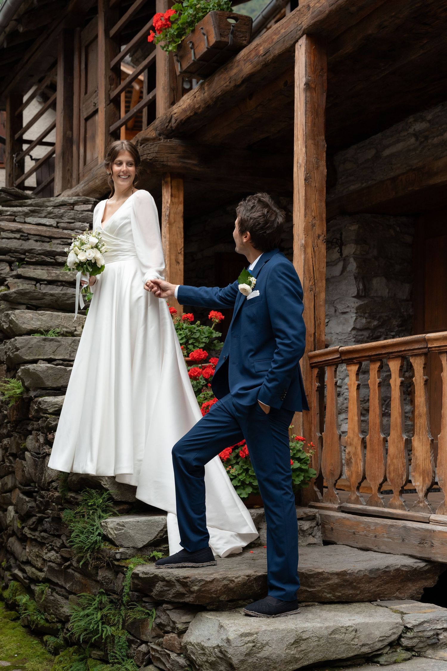 Smiling bride holds bouquet as groom leads her up stone steps of a rustic alpine chalet.