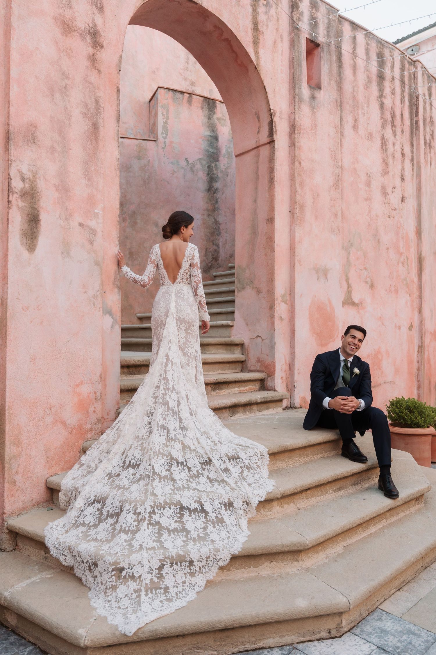 Elegant bride in backless lace wedding gown with long train poses on stone steps beside smiling seated groom.