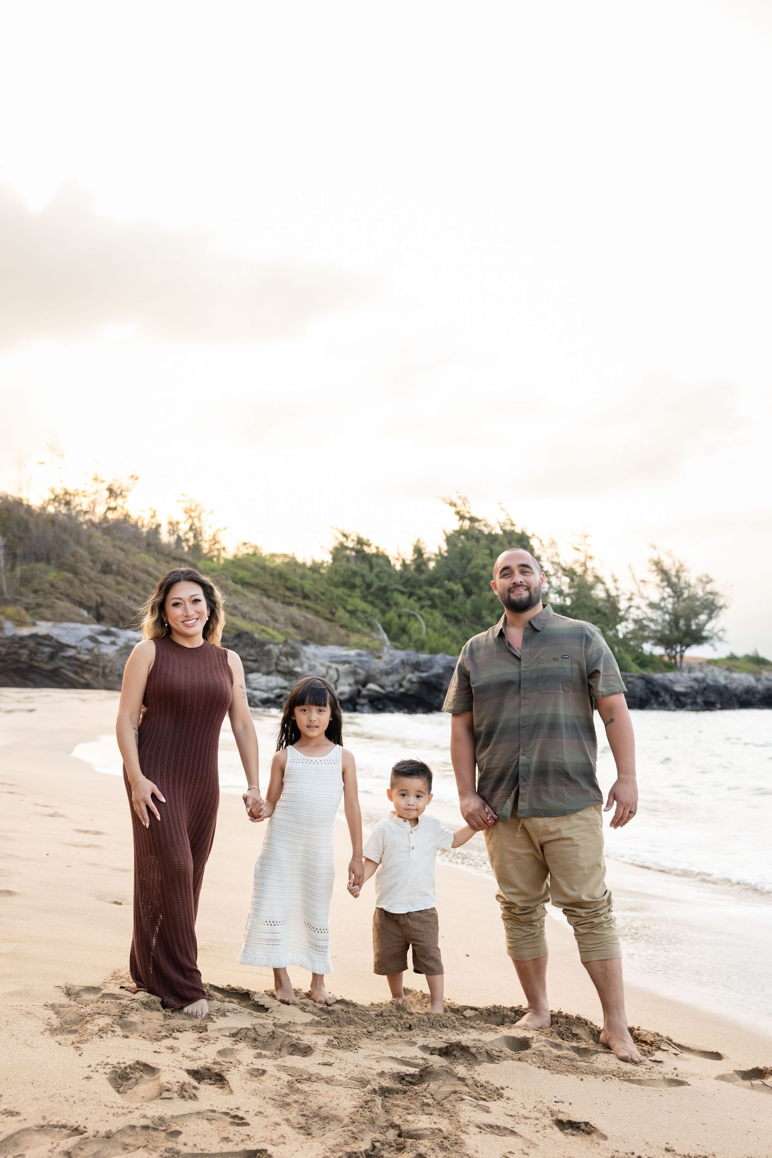 Family of four smiling together on a sandy Hawaiian beach during a beautiful golden hour sunset.