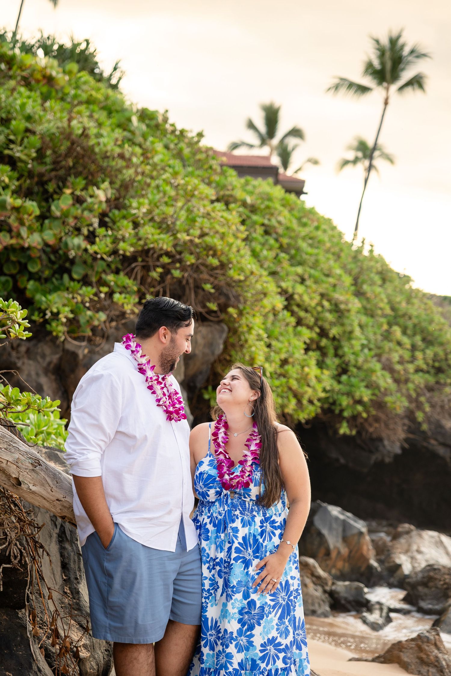 Couple wearing floral leis smiling at each other on a rocky Hawaiian beach with lush greenery.