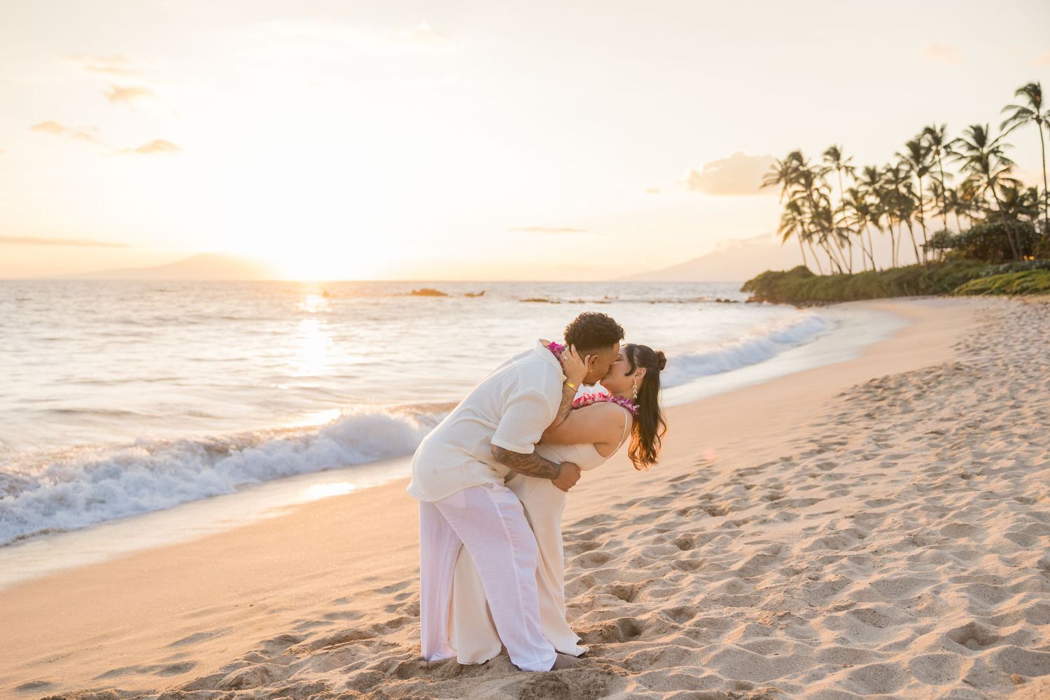 Couple sharing a romantic dip kiss on a sandy Hawaiian beach at golden sunset with palm trees and ocean waves.