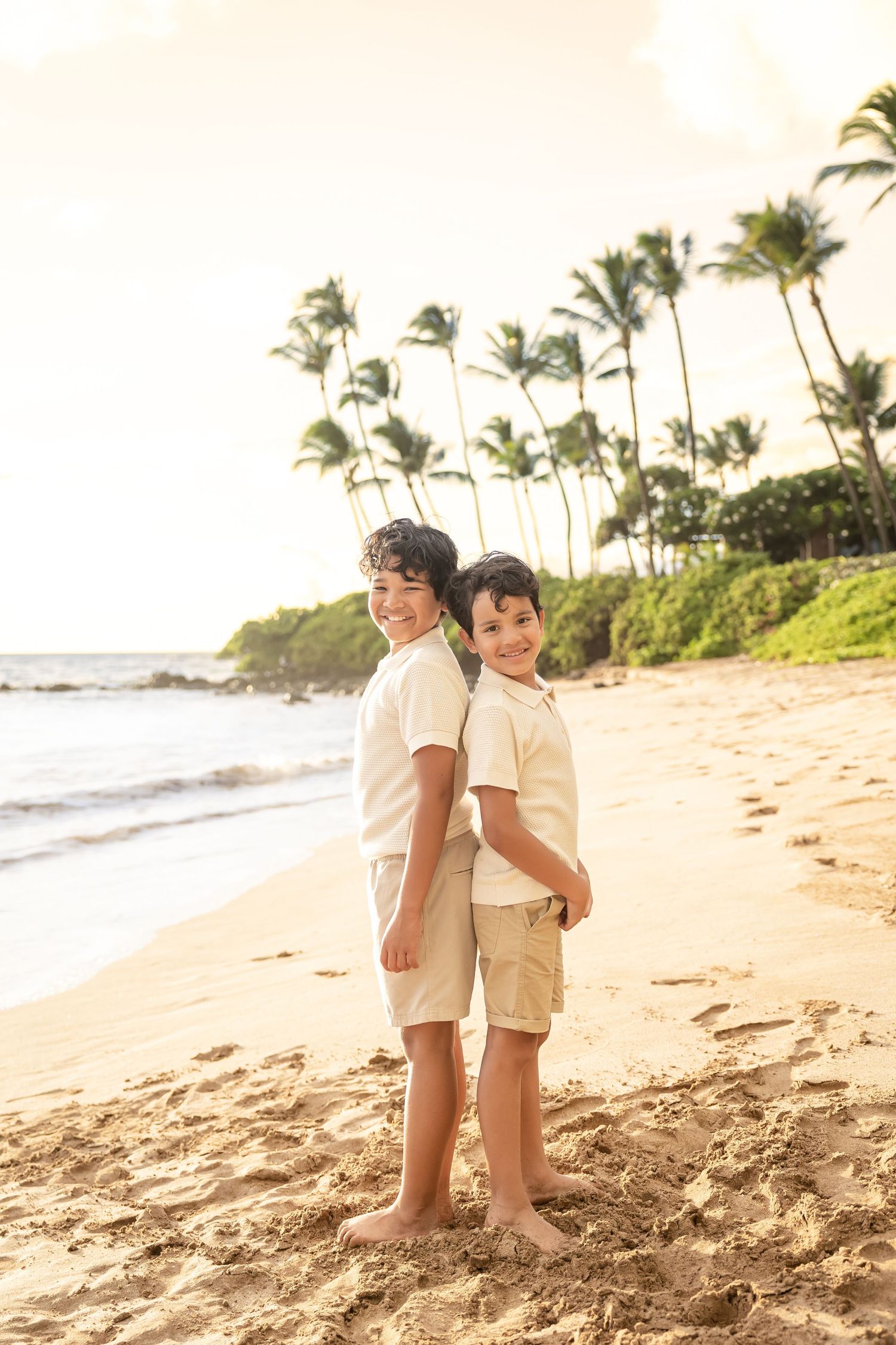 Two young brothers standing back to back on a Hawaii beach with palm trees.