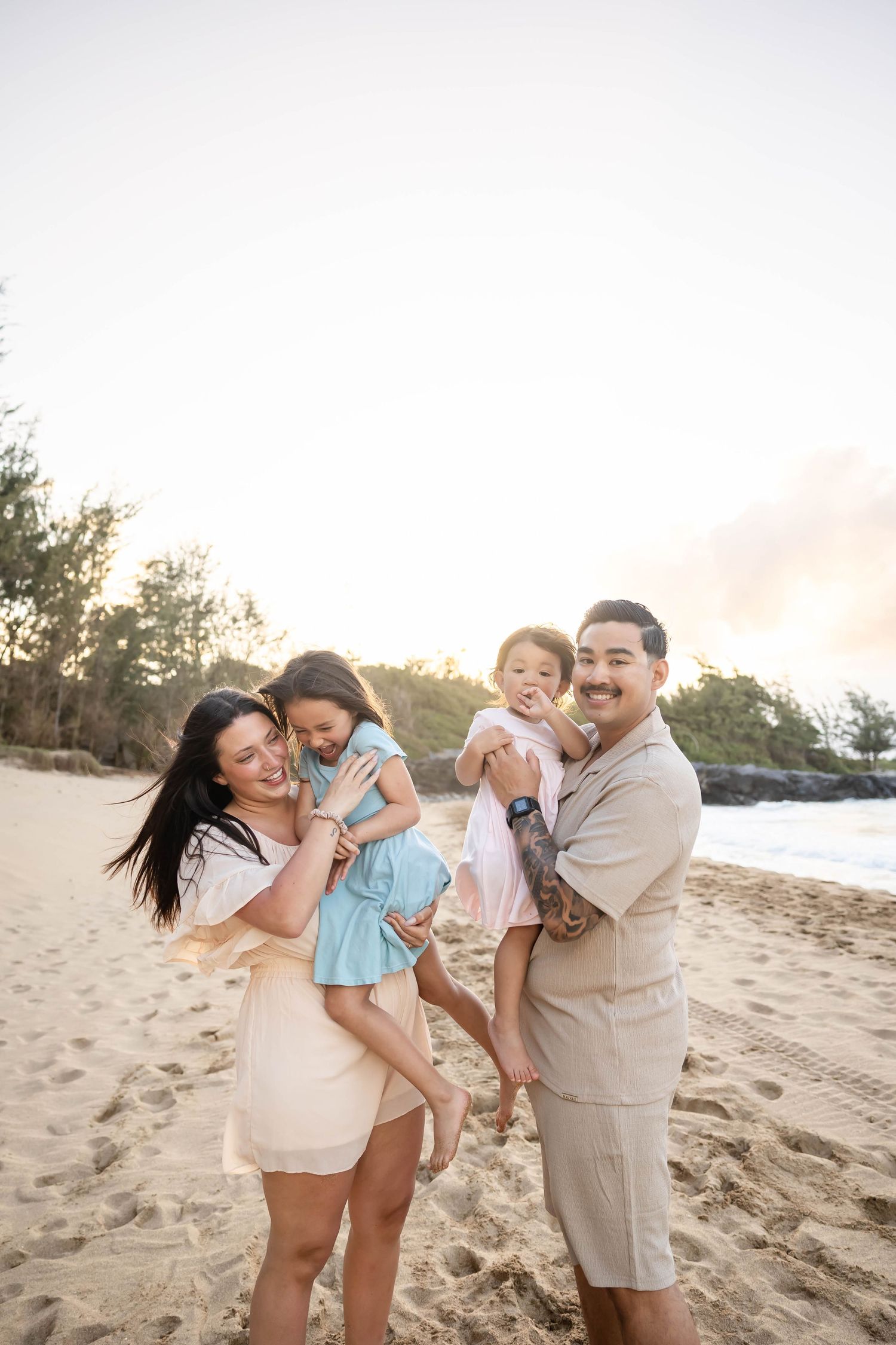 Joyful family of four posing on a beach at golden hour, parents holding their two daughters.