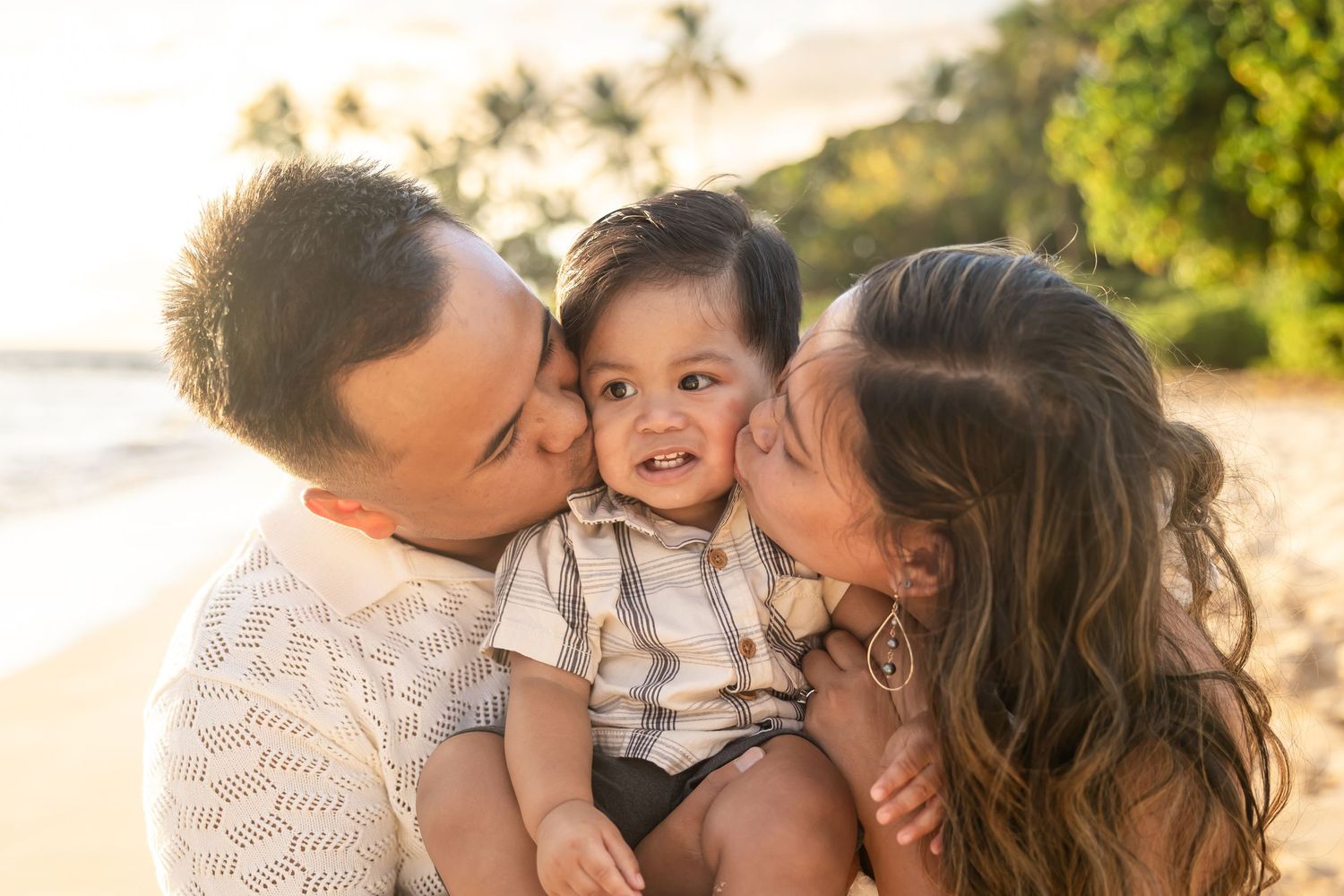 Two parents kissing their smiling baby boy on the cheeks on a sunny beach.