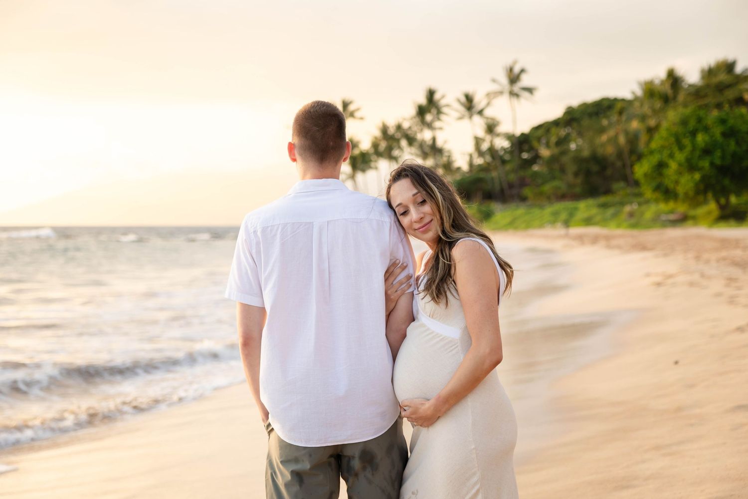 Pregnant woman in white dress leaning against partner on golden sandy beach with palm trees at sunset.