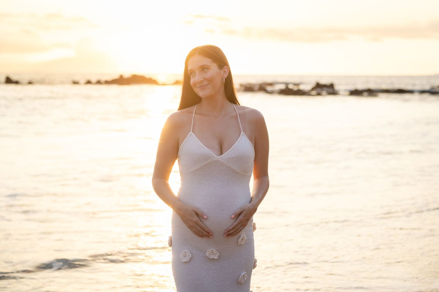 Pregnant woman in white floral dress smiling while holding baby bump on glowing sunset beach.