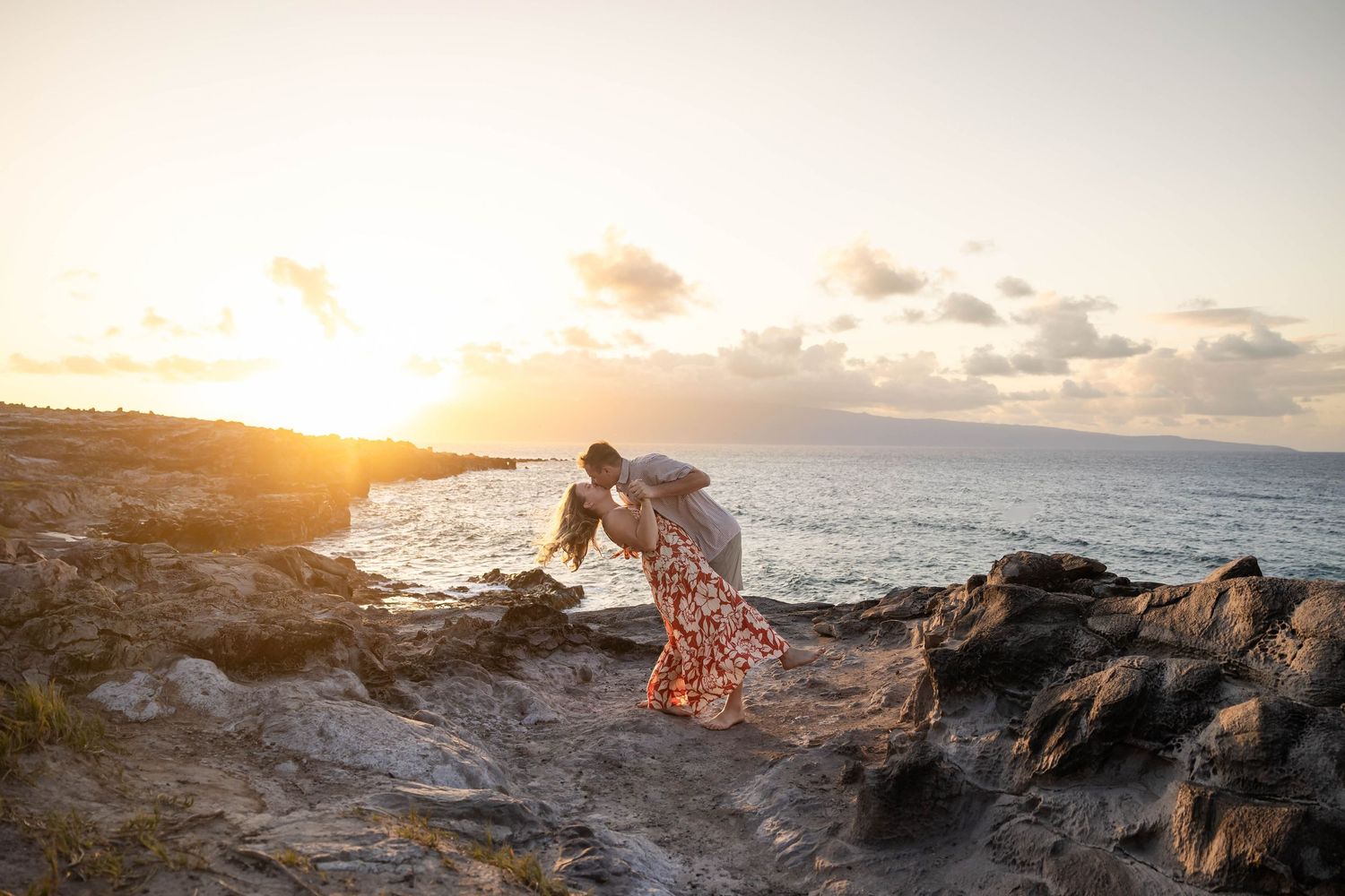 Couple sharing romantic dip kiss on rocky Hawaiian coastline at golden sunset.