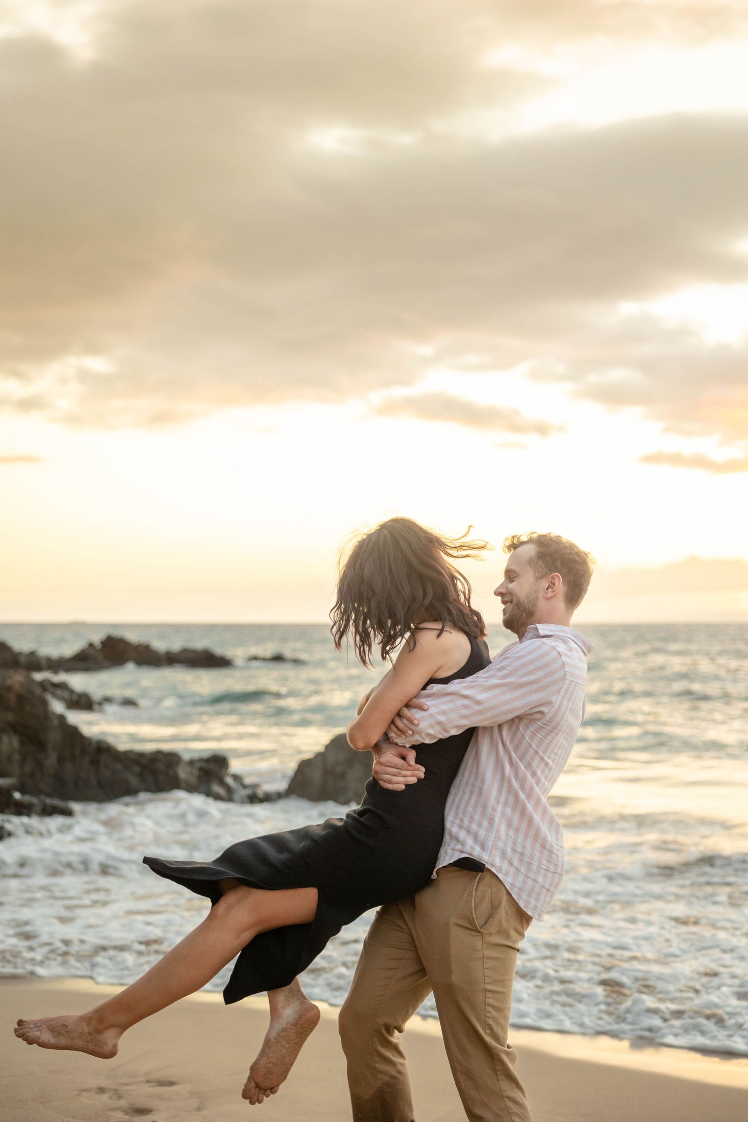 Man joyfully lifting woman in black dress on a sandy beach during a golden Hawaiian sunset.