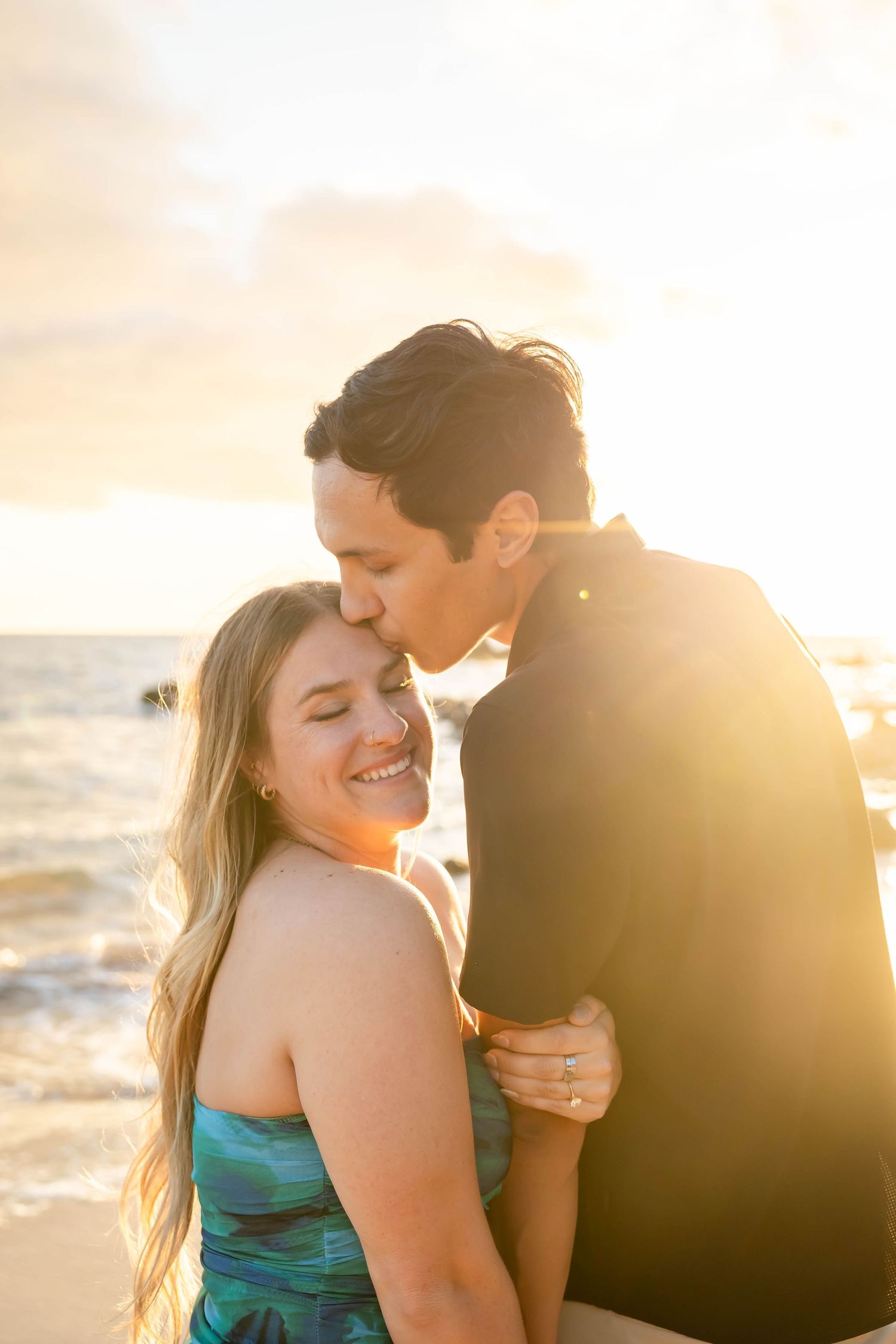 Couple sharing a romantic kiss on the beach at golden sunset, woman smiling with engagement ring visible.