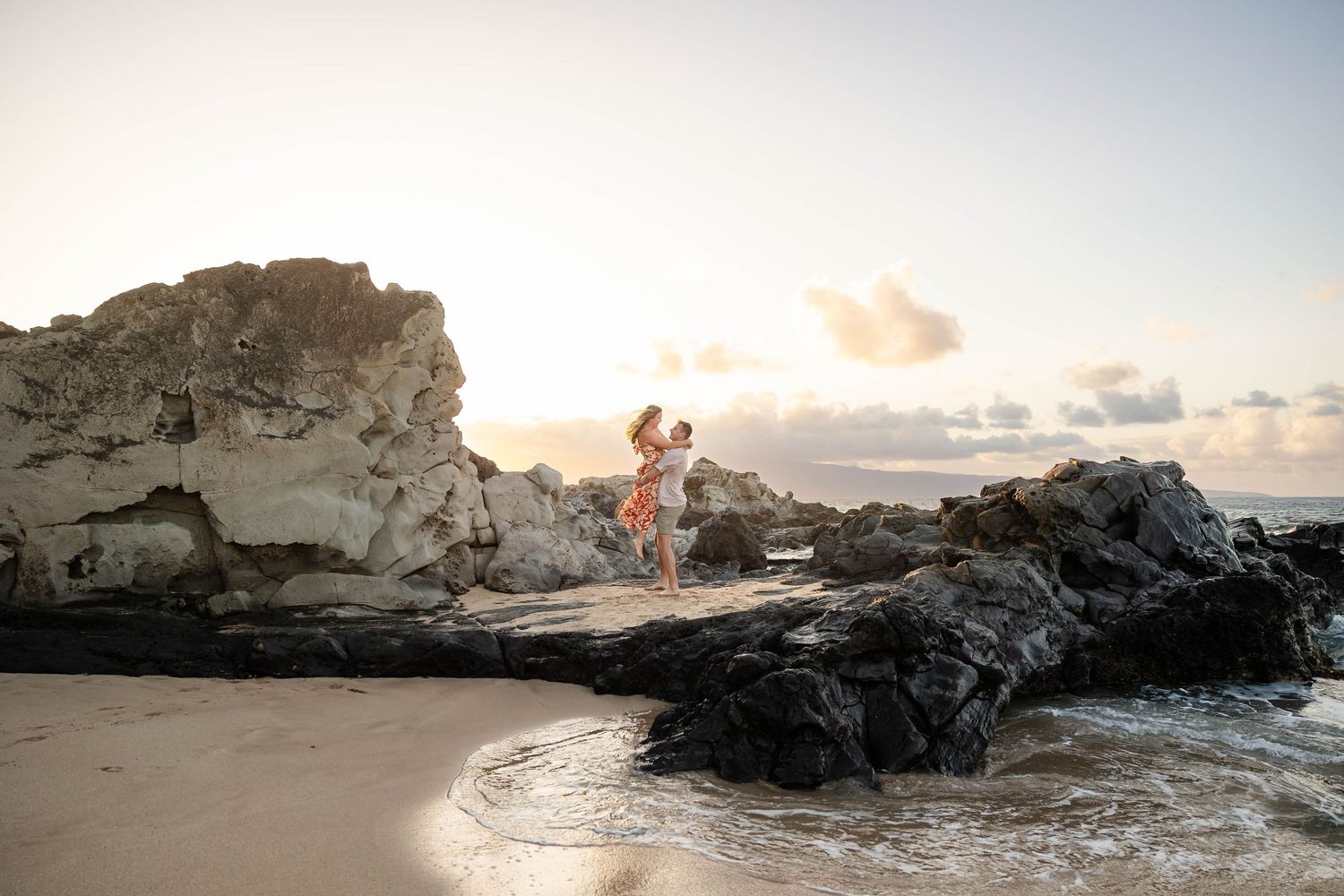 Couple embracing on volcanic rocks at a Hawaii beach during a stunning golden hour sunset.