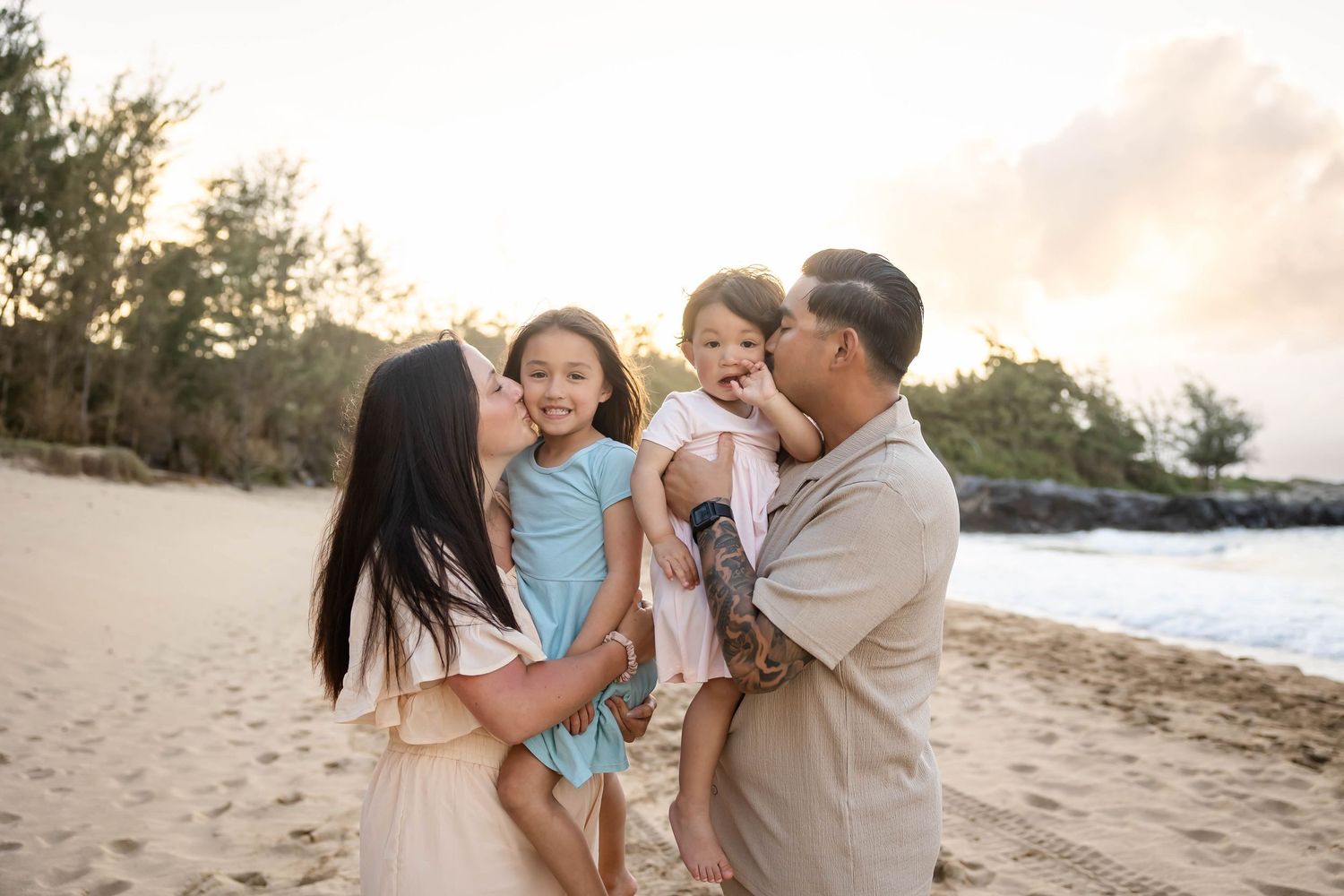 Loving parents kissing their two young daughters on a sandy beach at golden hour sunset.