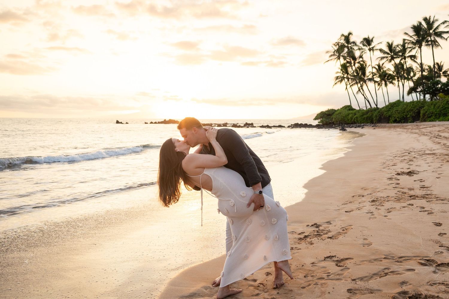 Couple sharing romantic dip kiss on sandy Hawaiian beach during golden hour sunset.