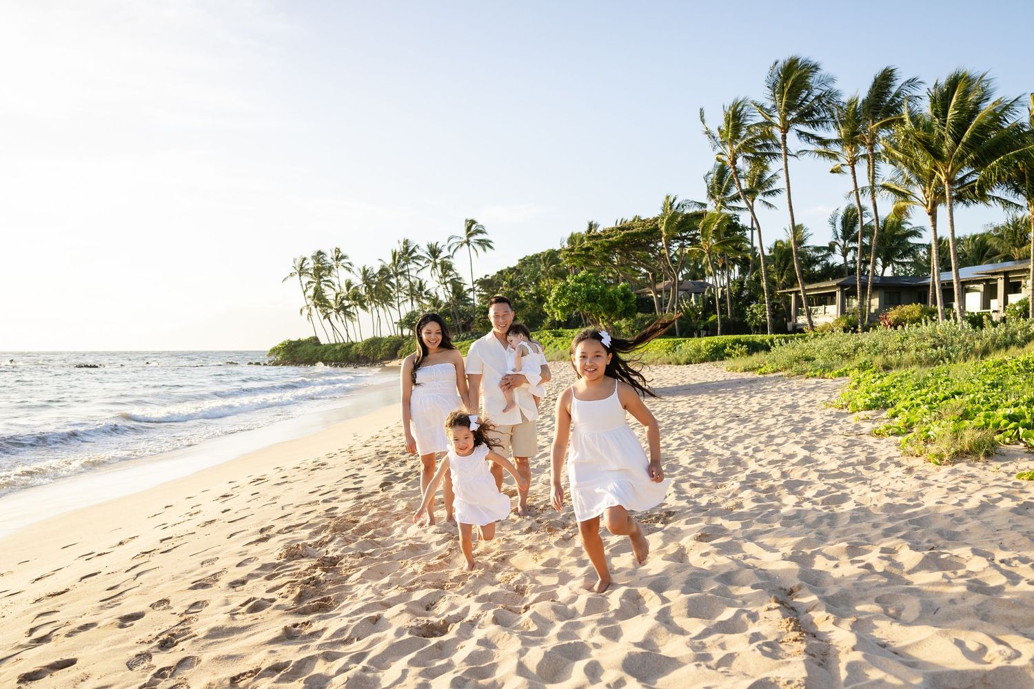 Happy family in white outfits running on a sunny tropical beach with palm trees in the background.