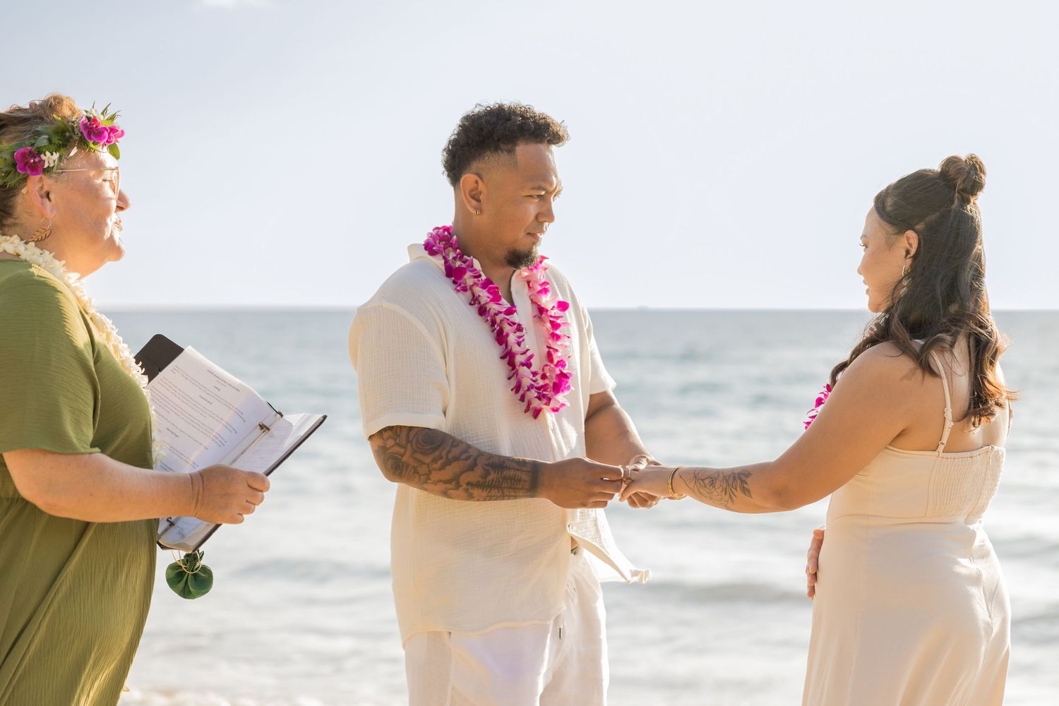 Couple exchanges vows on a Hawaiian beach with an officiant, wearing leis, with the ocean in the background.