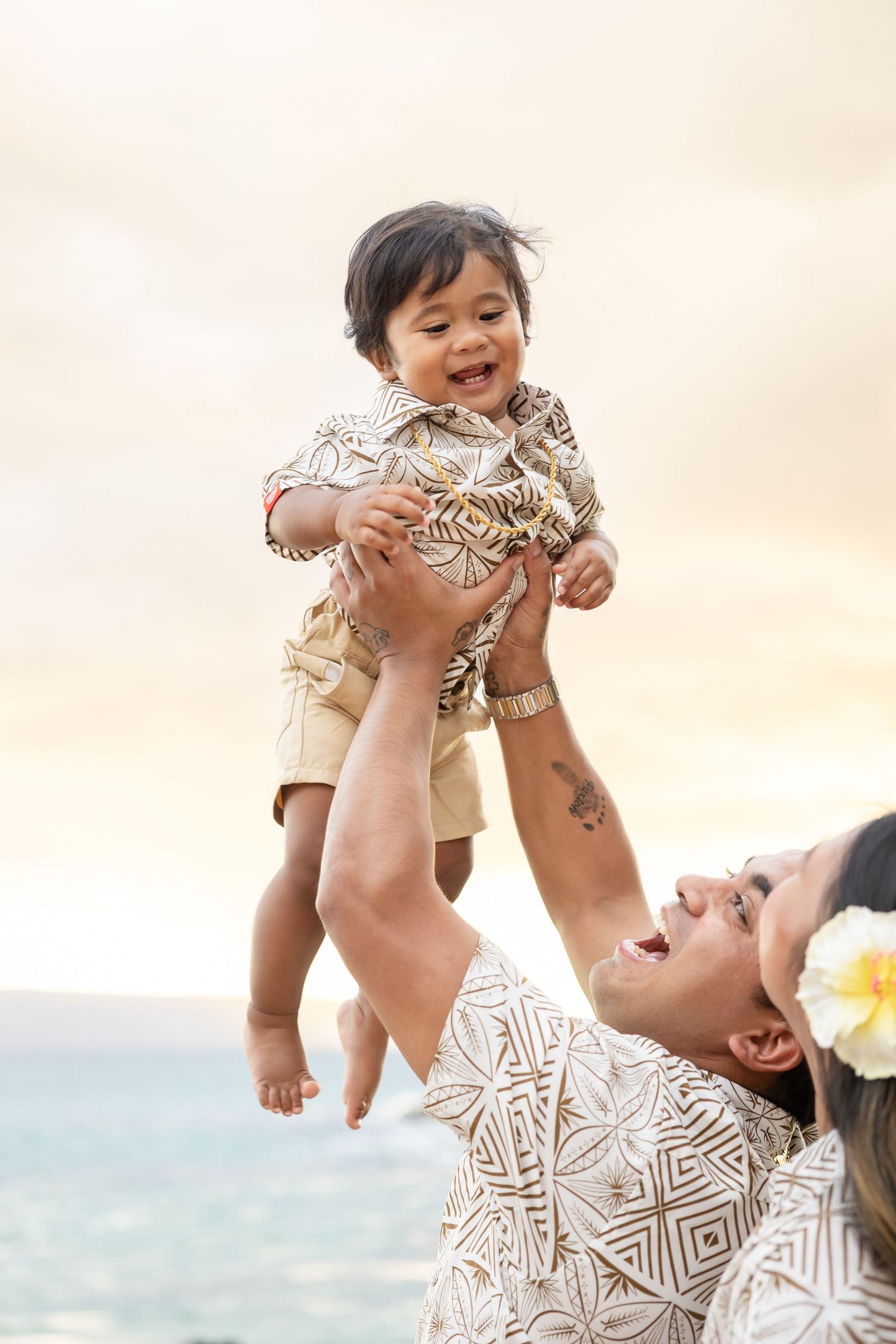 Father lifting laughing baby overhead on Hawaiian beach at sunset as mother watches joyfully.