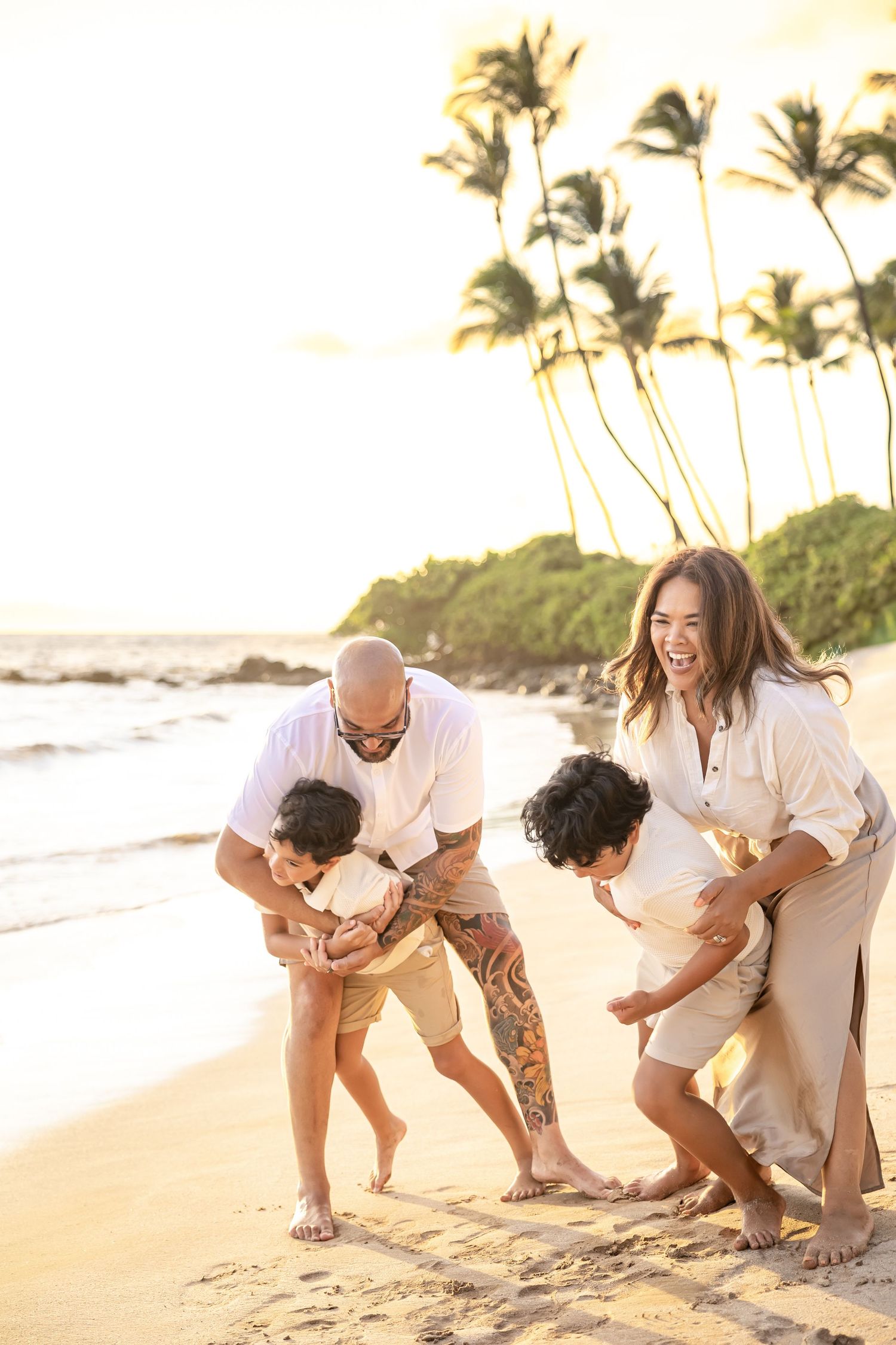 Family laughing and playing on a sandy Hawaiian beach at golden sunset with palm trees in background.