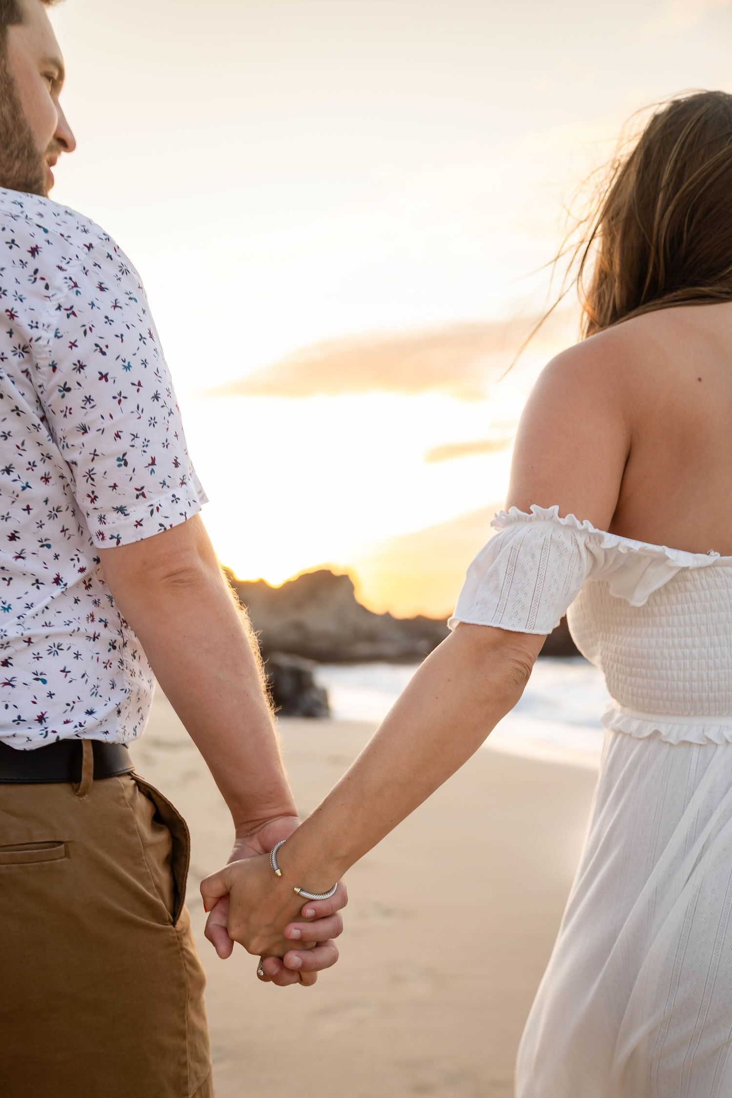 Close-up of couple holding hands on beach at sunset, woman in white off-shoulder dress with bracelet.