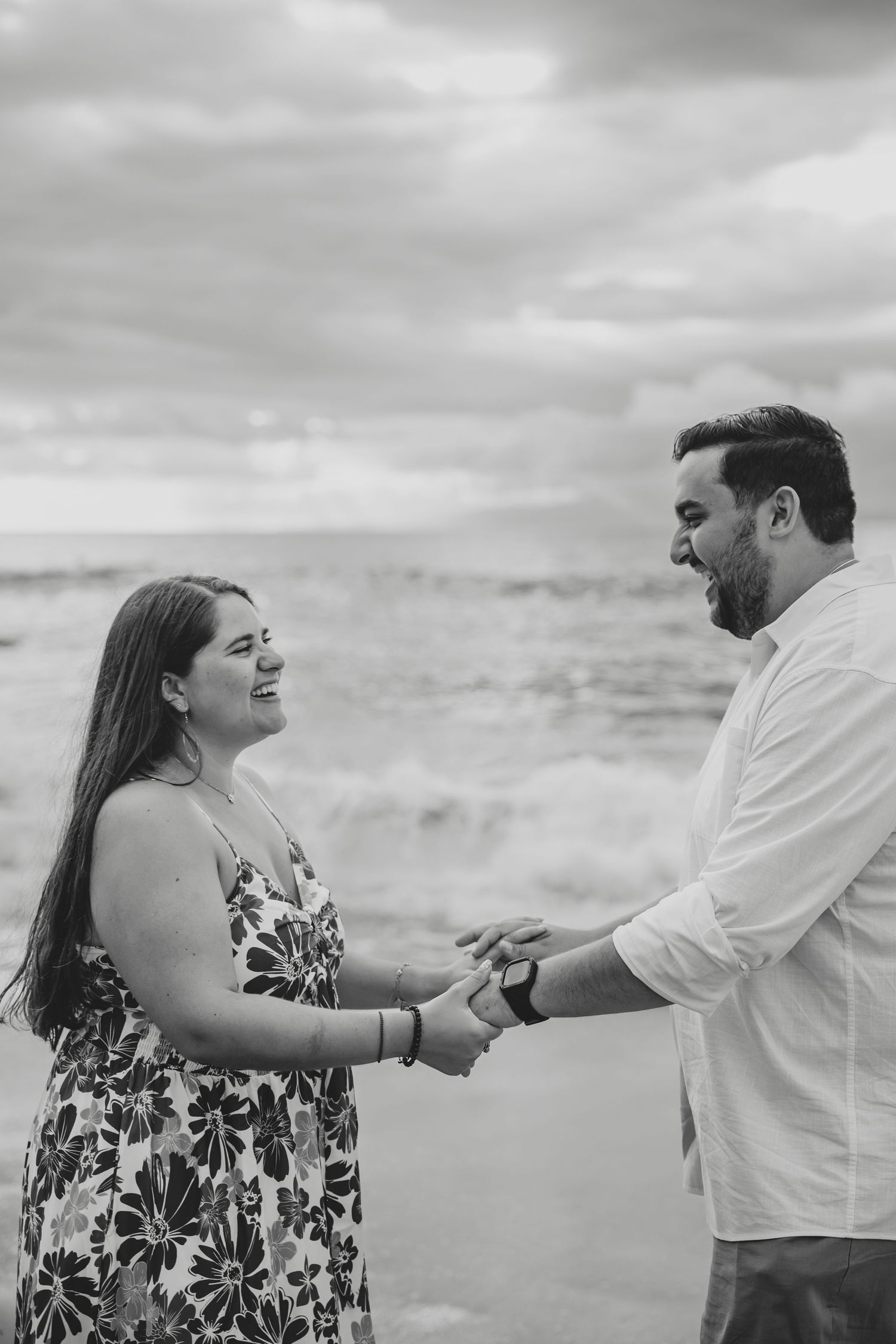 Black and white photo of couple holding hands and smiling at each other on a beach with ocean waves.