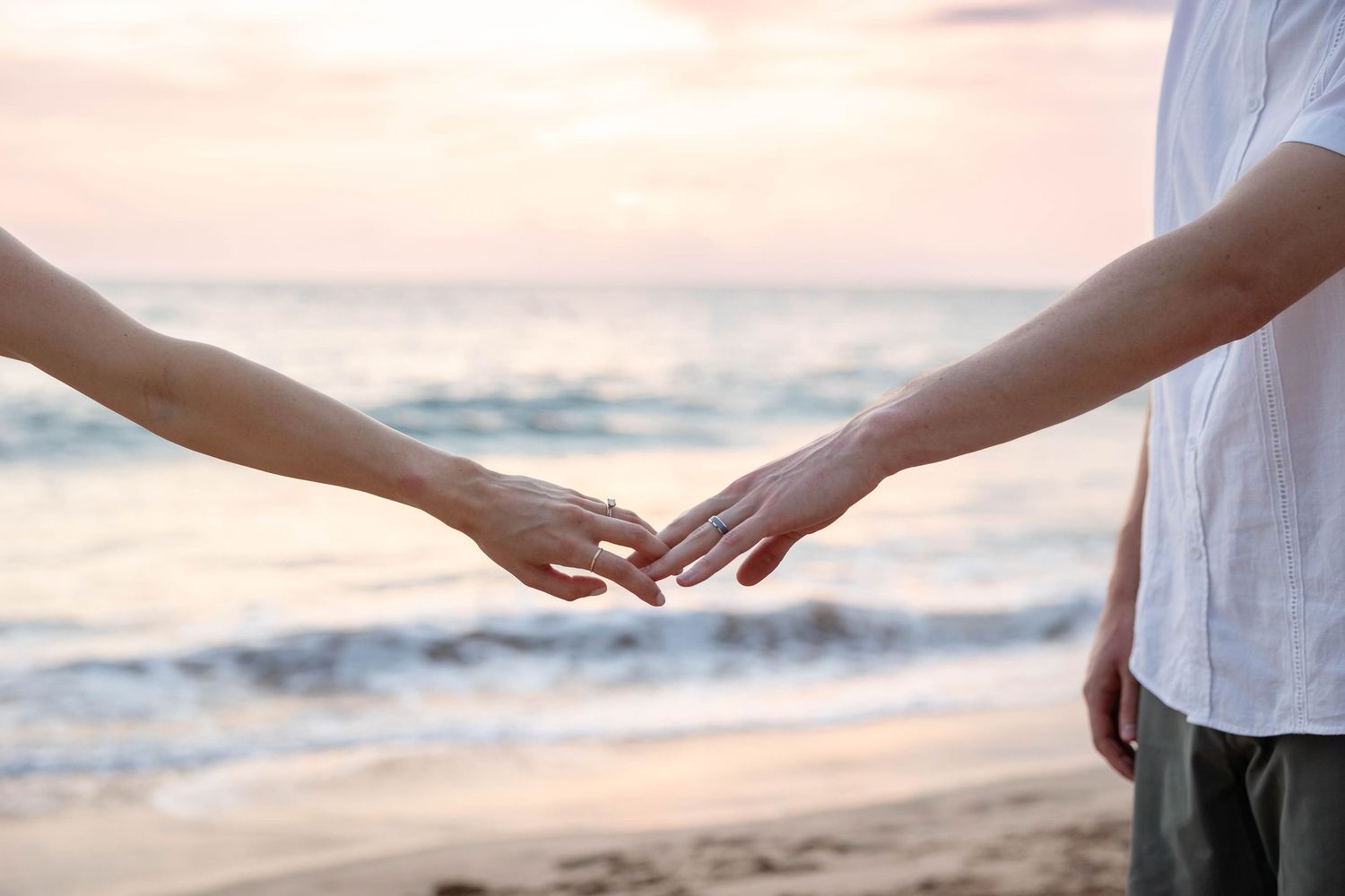 Close-up of couple gently touching fingertips on beach at sunset with ocean waves in background.