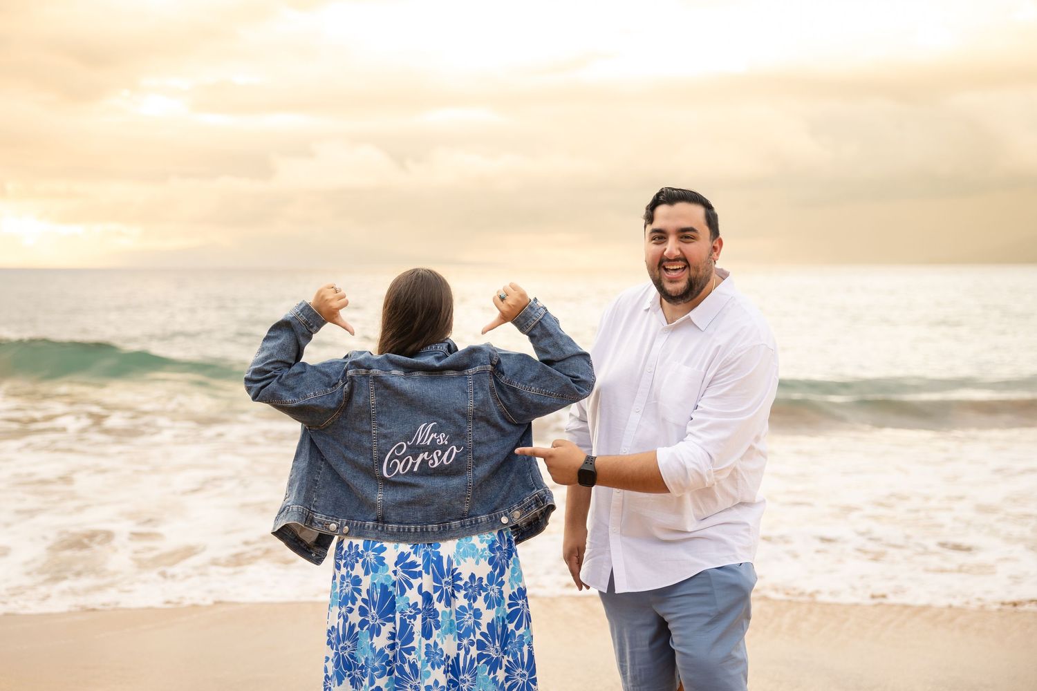 Excited woman showing off custom Mrs. Corso denim jacket to smiling fiance on Hawaiian beach at sunset.