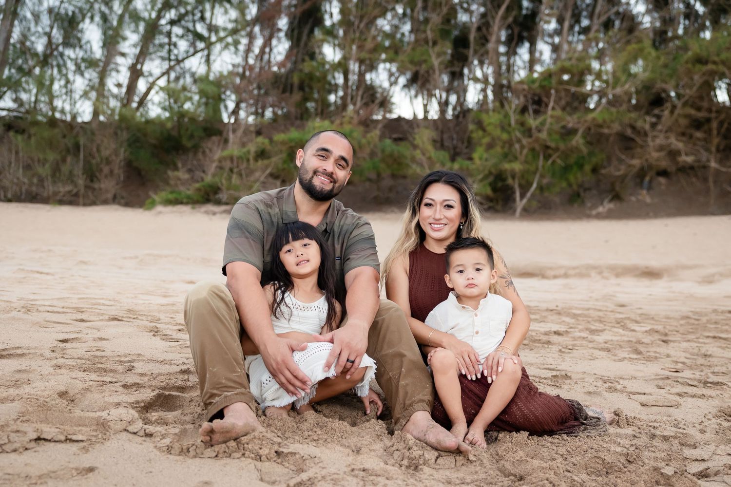 Happy family of four sitting together on sandy Hawaiian beach surrounded by lush tropical trees.