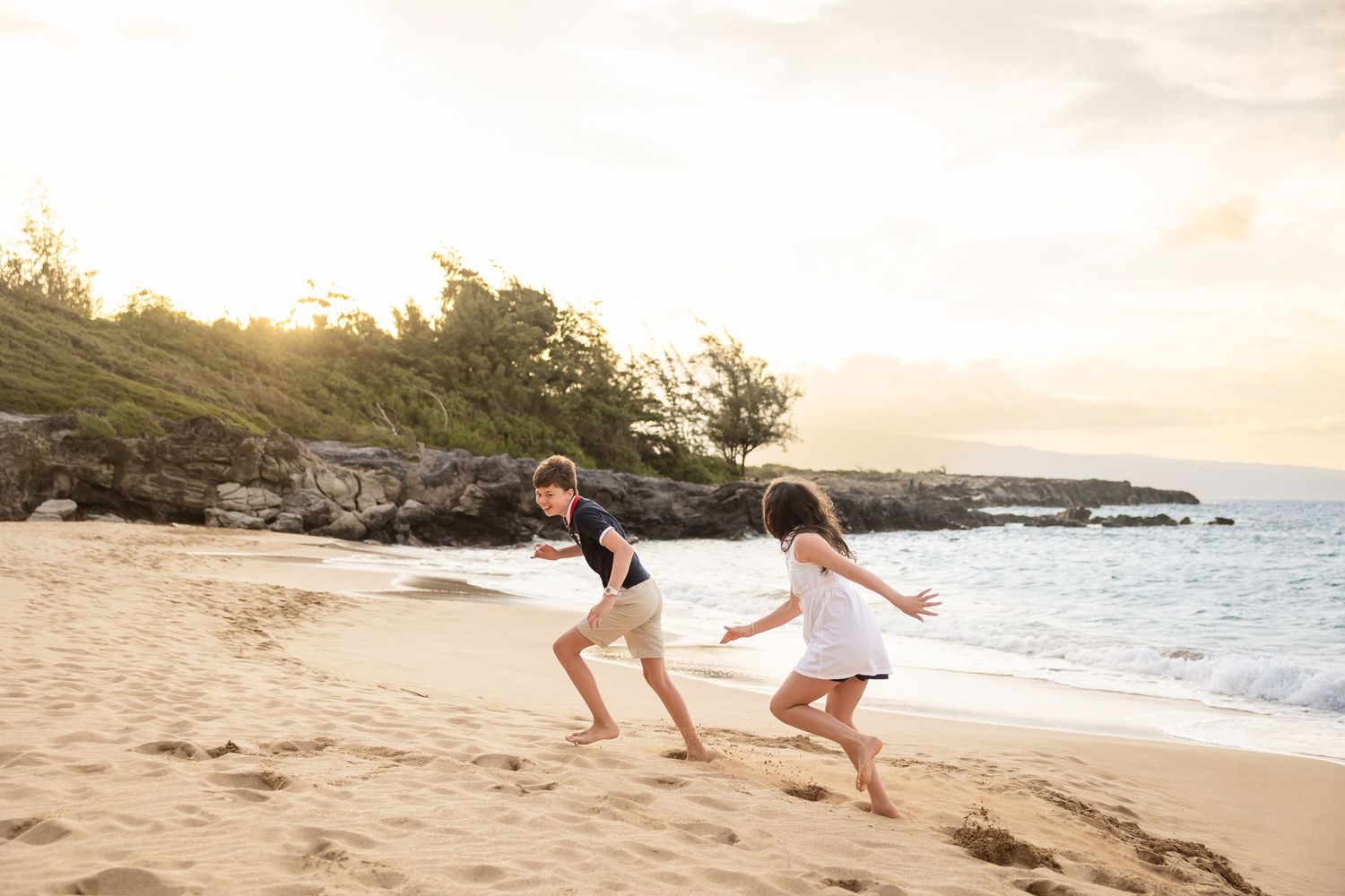 Two children running and playing on a sandy Hawaiian beach at golden sunset near the ocean.