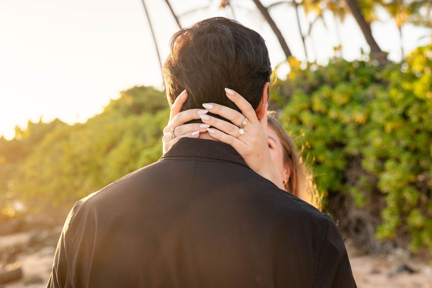 Bride showing off her engagement ring while embracing her partner near tropical greenery at sunset.