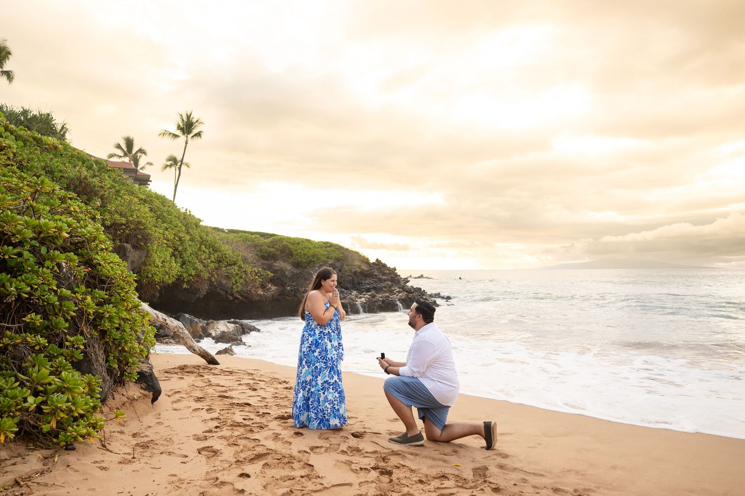 Man proposing on one knee to woman in blue floral dress on sandy Hawaiian beach at sunset.