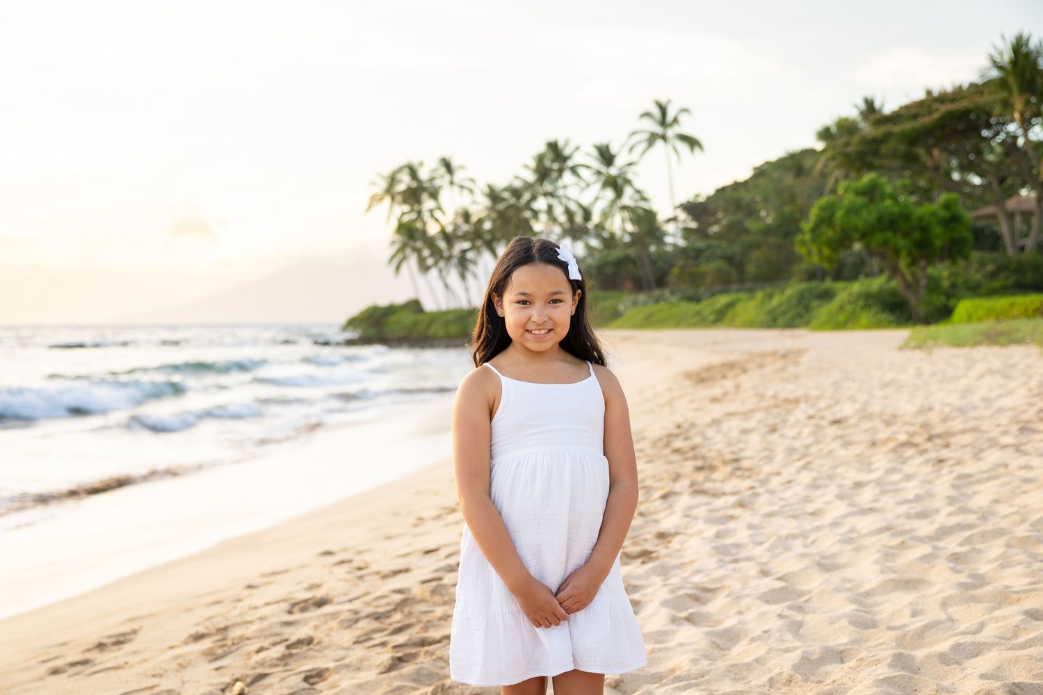 Young girl in white dress smiling on sandy Hawaiian beach with palm trees and ocean waves at sunset.