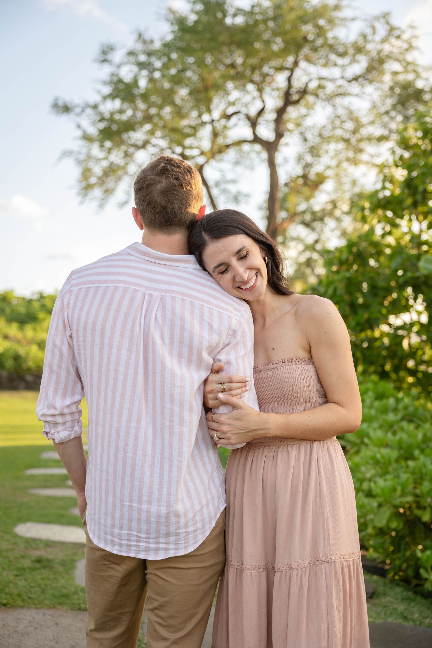 Smiling woman in pink dress embracing her partner from behind in a lush tropical garden setting.