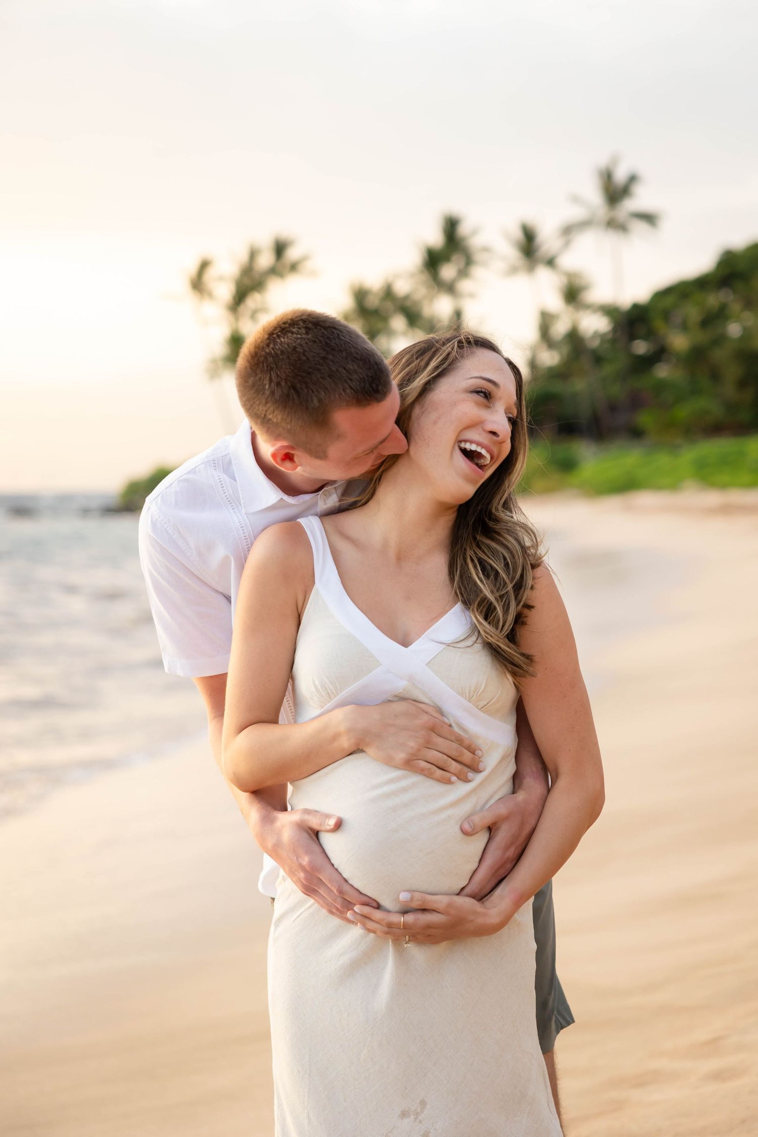 Couple sharing joyful moment on tropical beach at sunset, man kissing pregnant woman's cheek as she laughs.