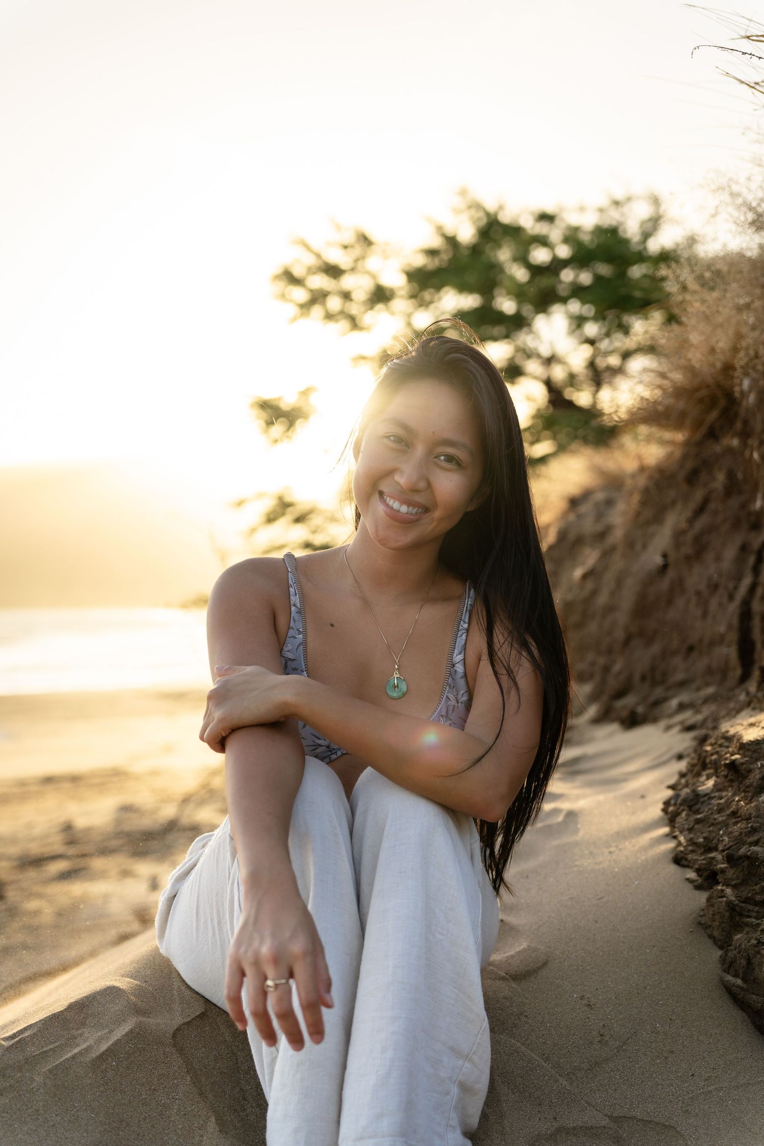 Smiling young woman with long dark hair sits on a sandy beach at golden hour, wearing white pants and a floral top.