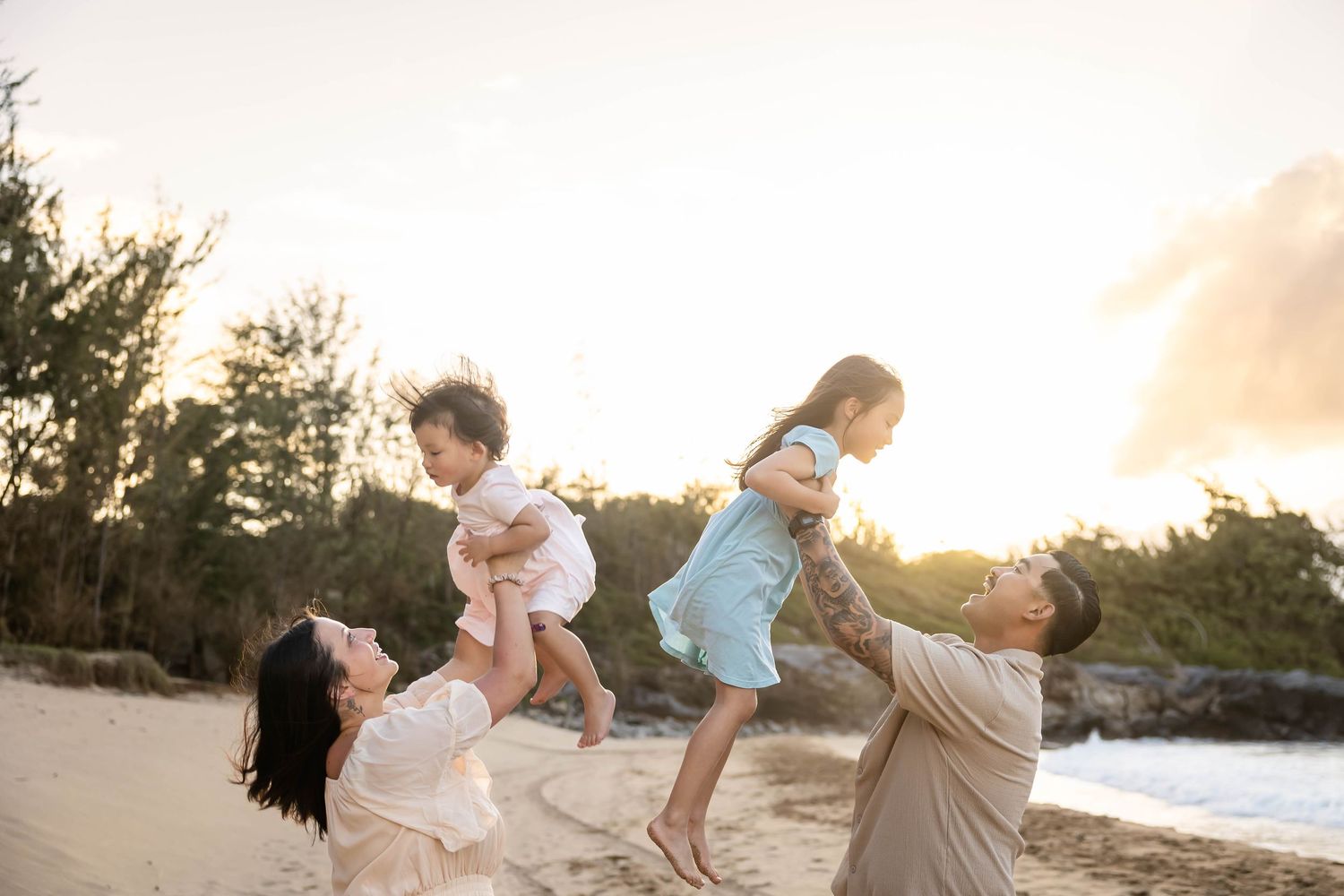 Happy family playing on beach at sunset, parents lifting two young children in the air.