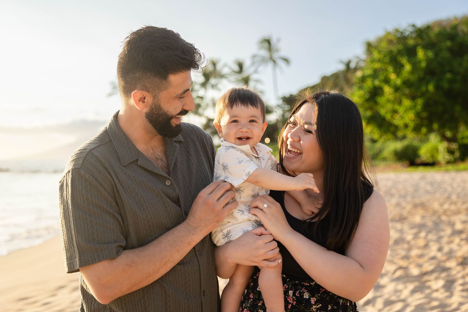 Happy young family with baby enjoying a sunny beach day in Hawaii with palm trees.