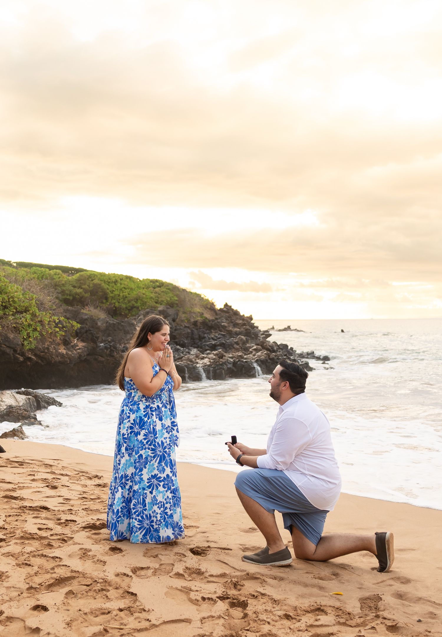 Surprise beach proposal at sunset in Maui, woman in blue floral dress reacts with joy.