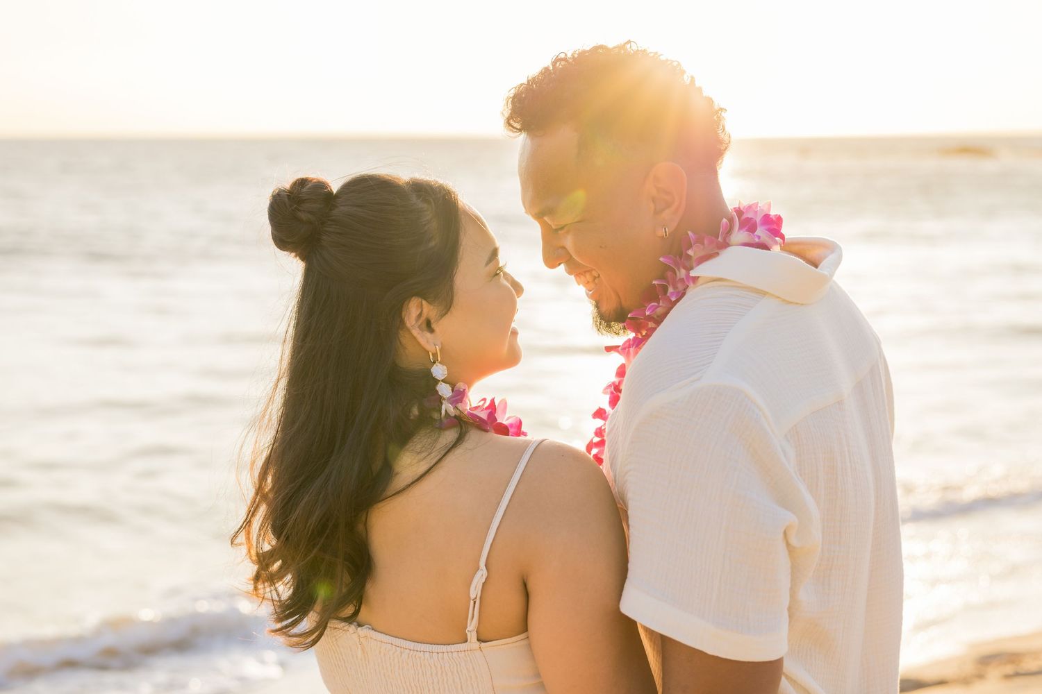 Couple sharing a romantic moment on a beach at sunset, woman in white dress, man wearing a pink lei in Hawaii.