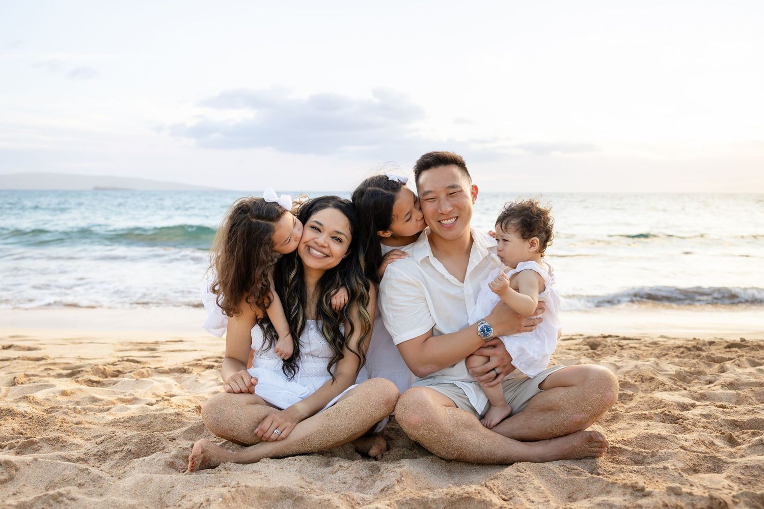 Happy Asian family of five smiling together on a sandy beach at sunset, parents with three young children.