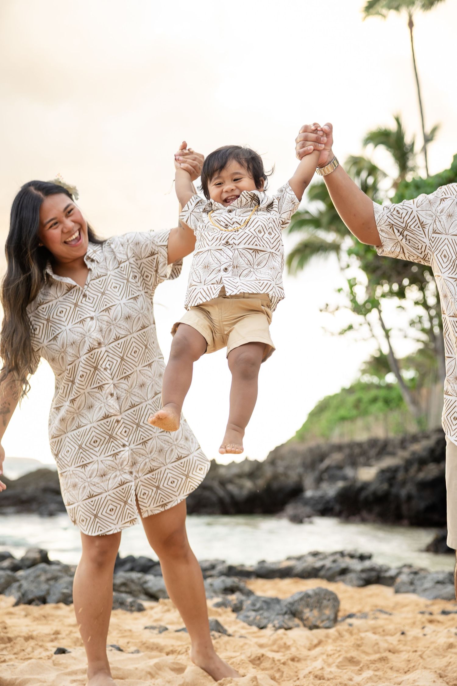 Parents swing their laughing toddler by the hands on a tropical Hawaiian beach at sunset, all wearing matching outfits.
