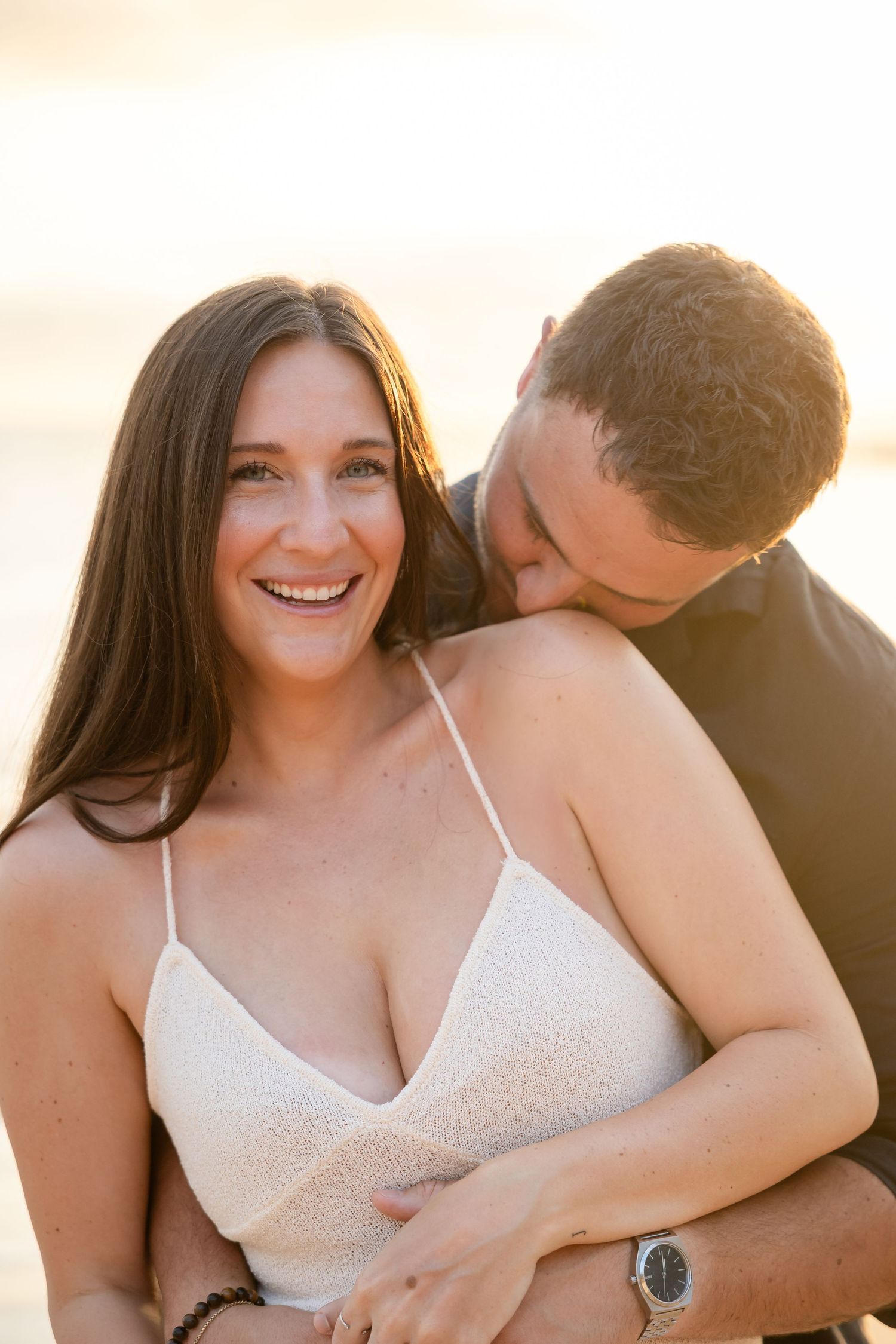 Smiling woman in white dress with man kissing her cheek during golden sunset beach photo session.