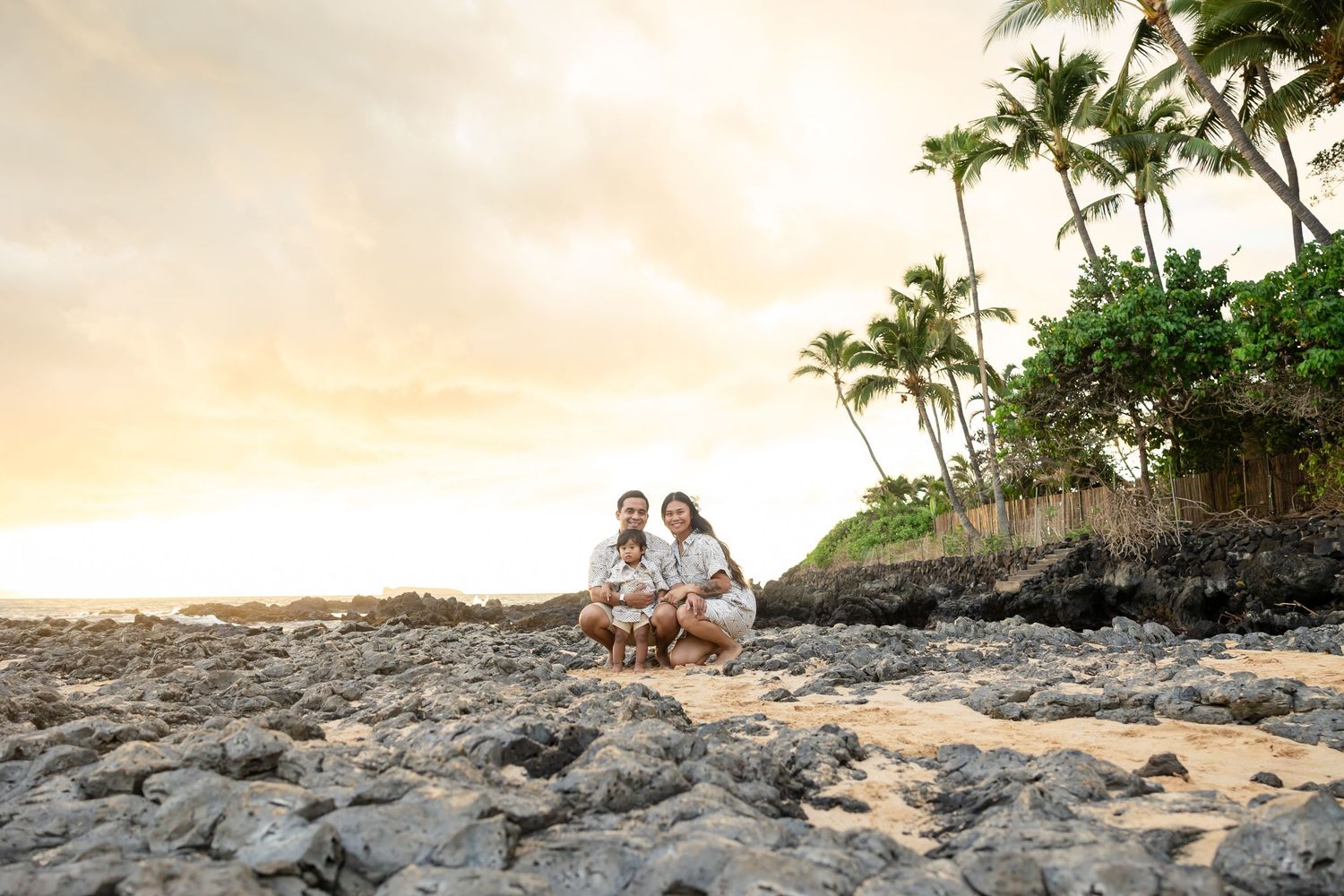 Family of three posing on rocky Hawaiian beach at sunset with palm trees and golden sky.