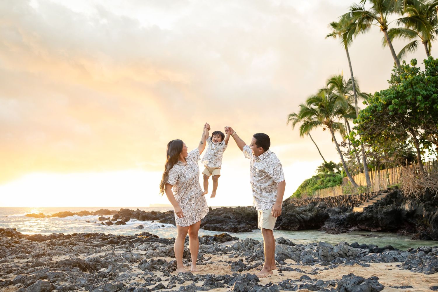 Family lifting young child between them on rocky Hawaiian beach at golden sunset with palm trees.
