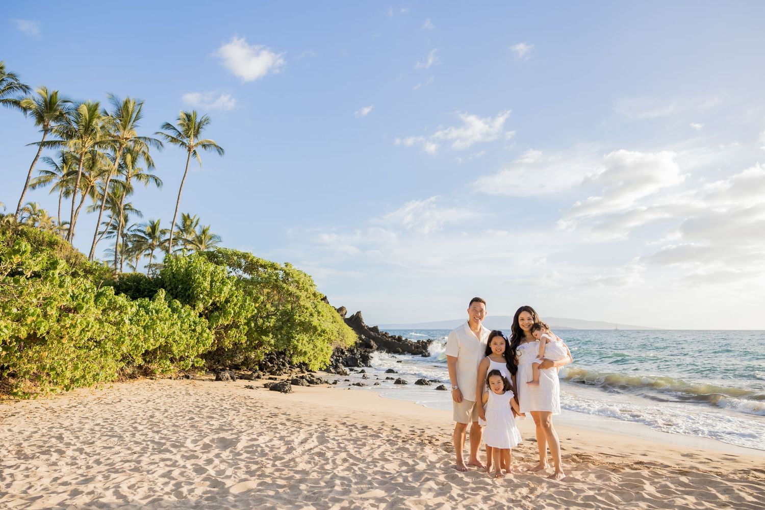 Family of four posing together on a sunny Hawaiian beach with palm trees and ocean waves in the background.