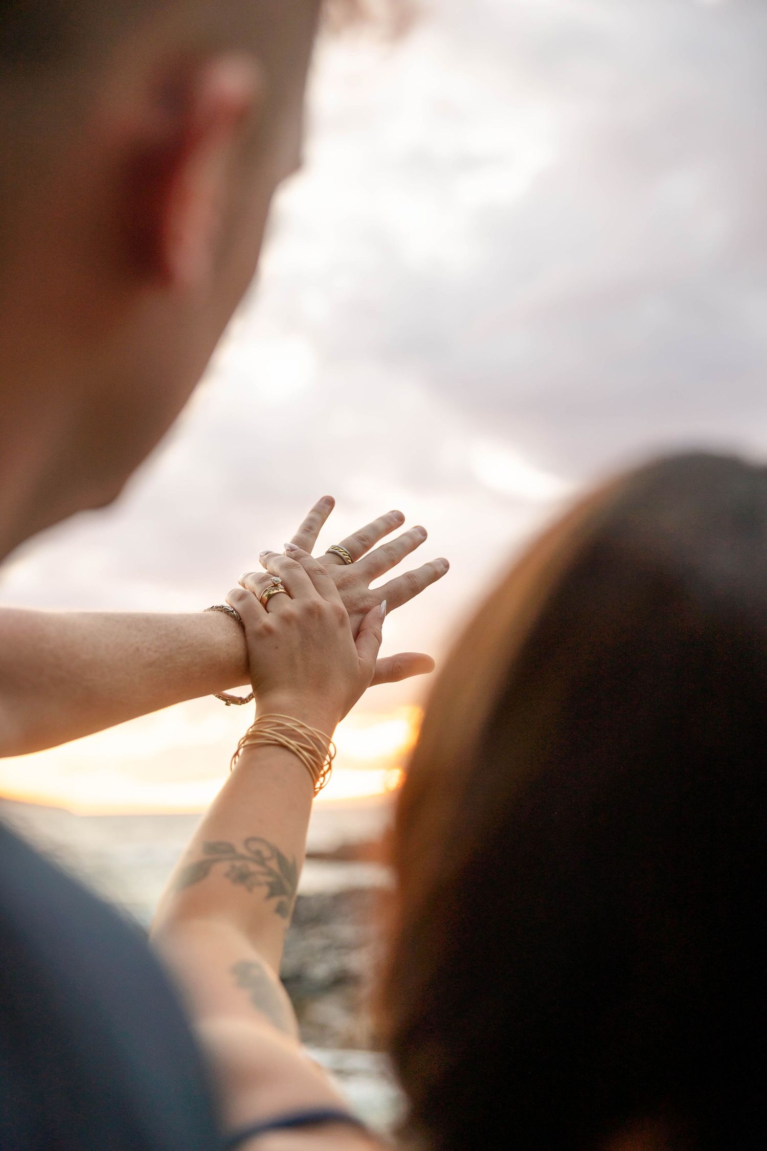 Close-up of couple holding hands displaying gold rings and bracelets with floral tattoo at sunset.