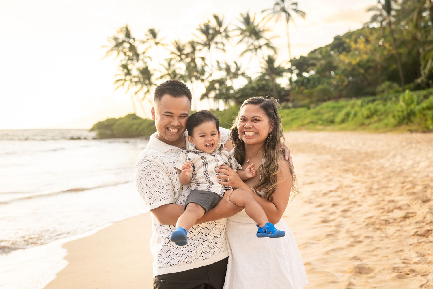 Happy young family with baby boy posing together on a sandy Hawaiian beach at sunset.