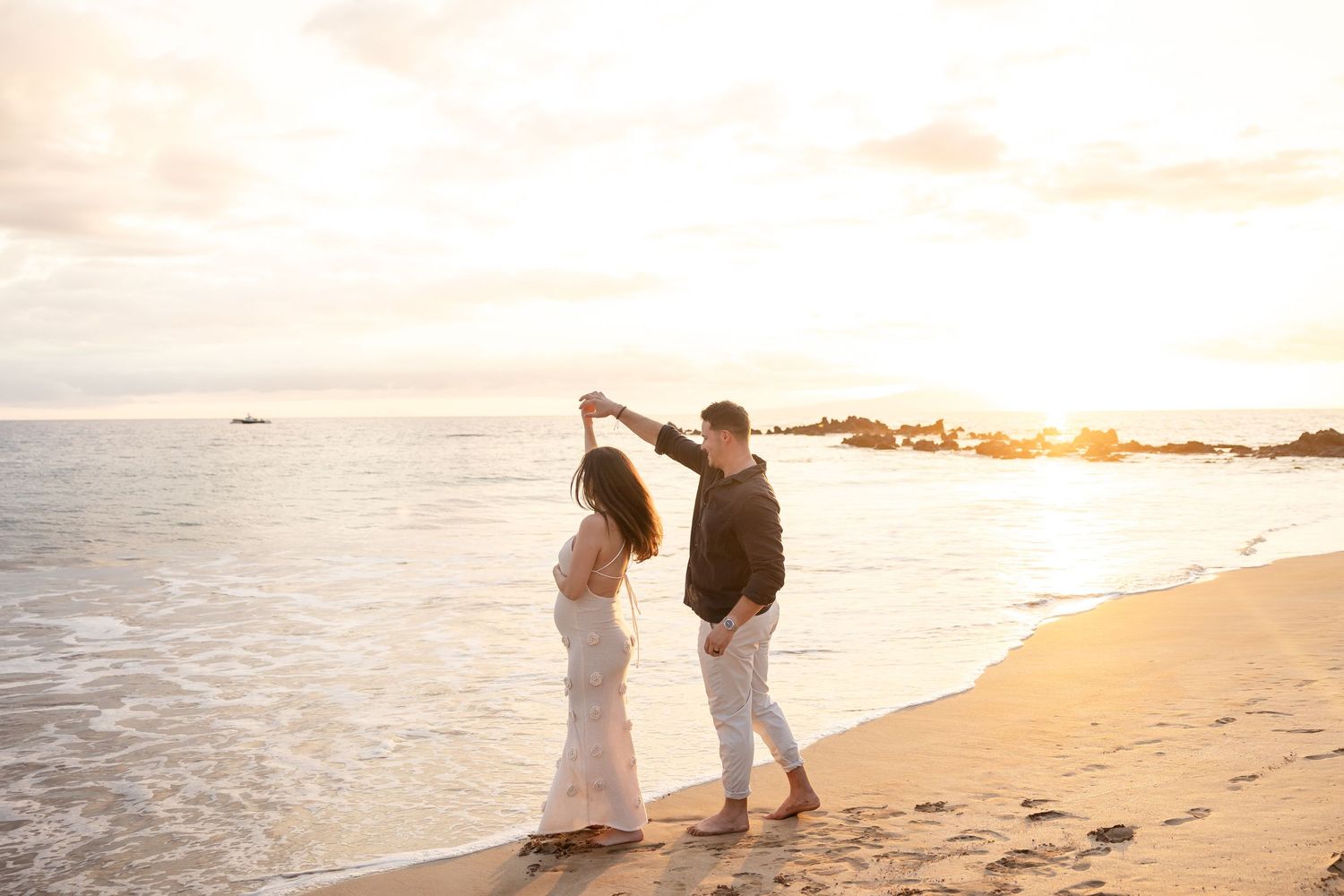 Man twirling woman in floral dress on sandy beach during beautiful golden sunset.
