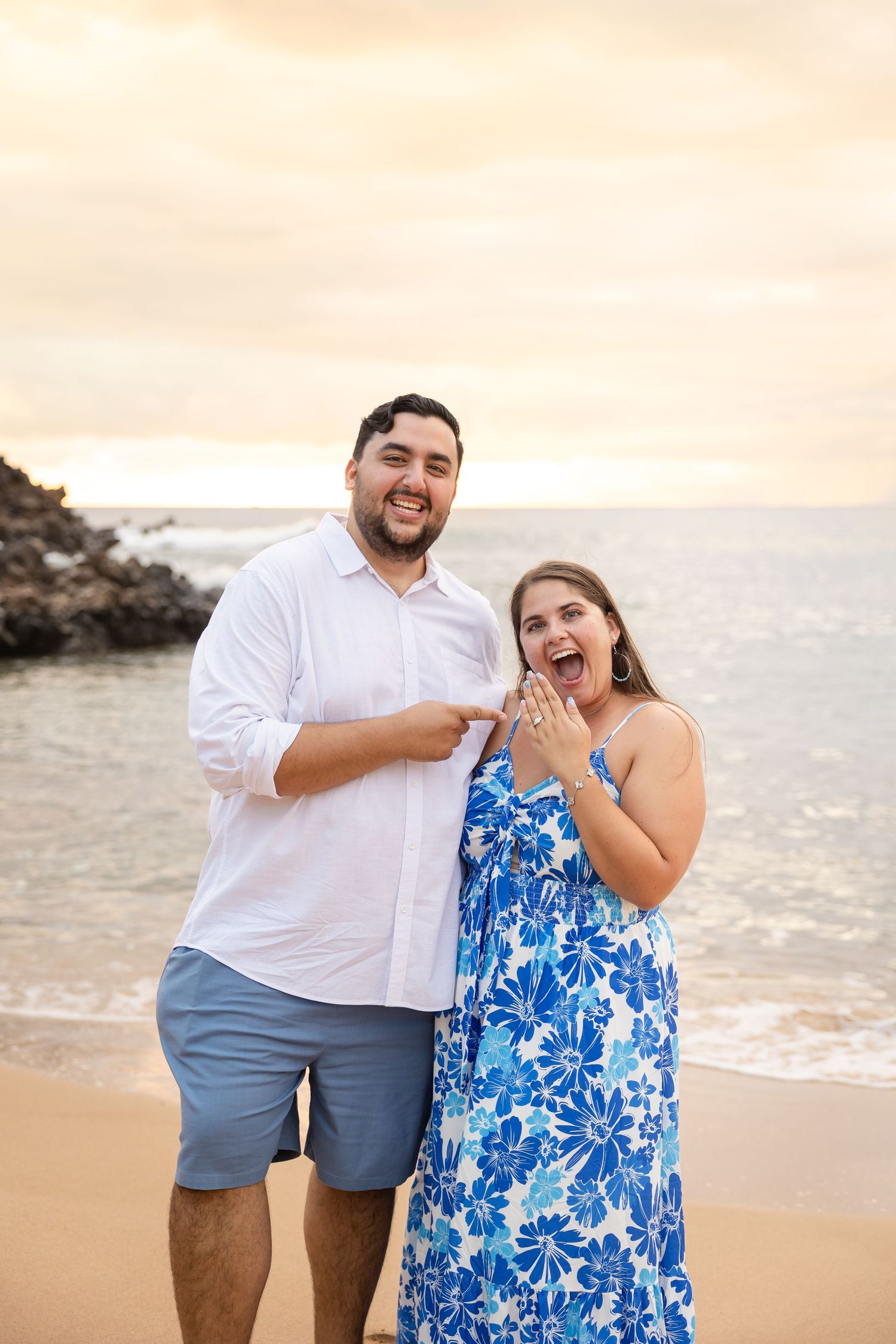 Excited newly engaged couple showing off engagement ring on a Hawaiian beach at golden sunset.