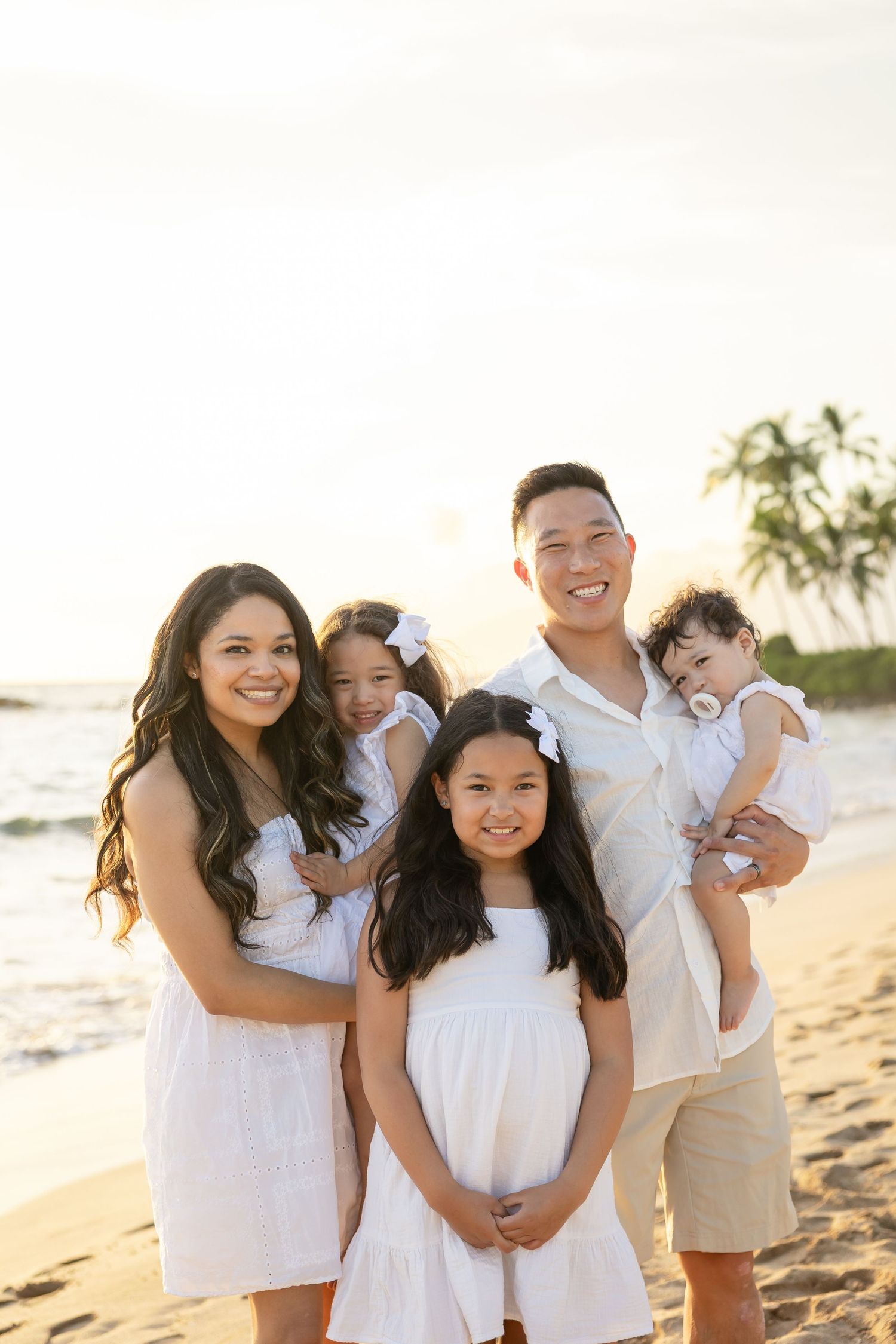 Family of five in white outfits posing on a sunny beach with palm trees, smiling together for a portrait.